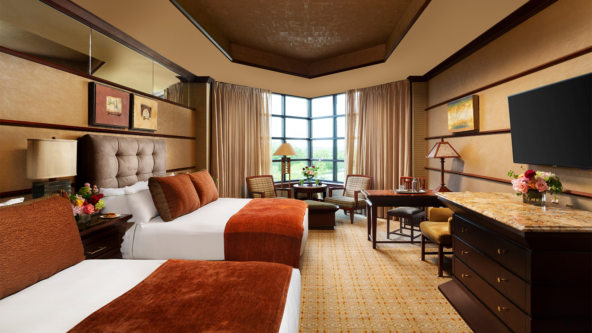 interior shot of Falling Rock's luxury double room. There are two queen size beds on the left side of the room. They have plush headboards and are decorated with white and burnt orange bedding. On the right side of the room is a dining area and a dresser with a flat screen TV mounted above it. There are windows overlooking the resort grounds with a sitting area.