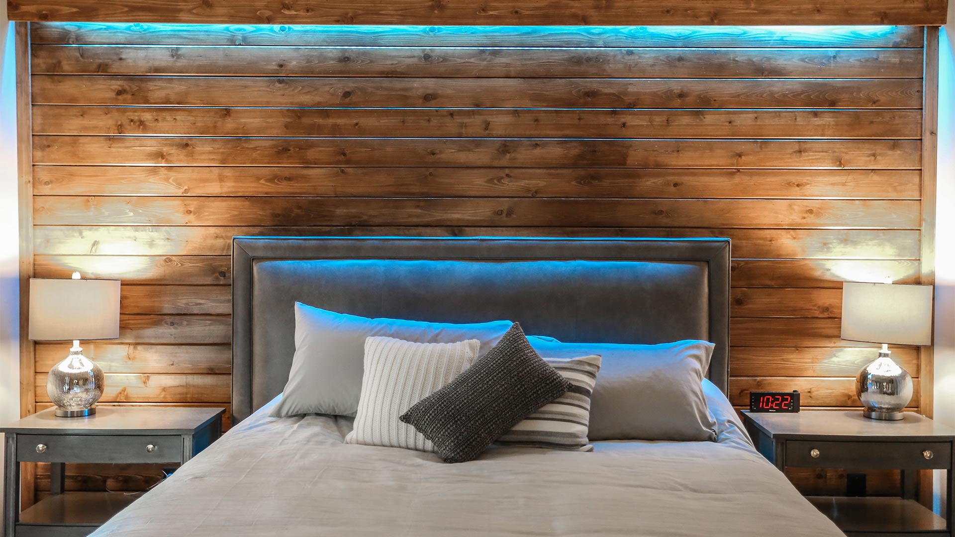 detail shot of a bedroom. There is calming blue mood lighting above the bed. The bed is neutral tones of gray. The wall behind the bed is a dark wood.