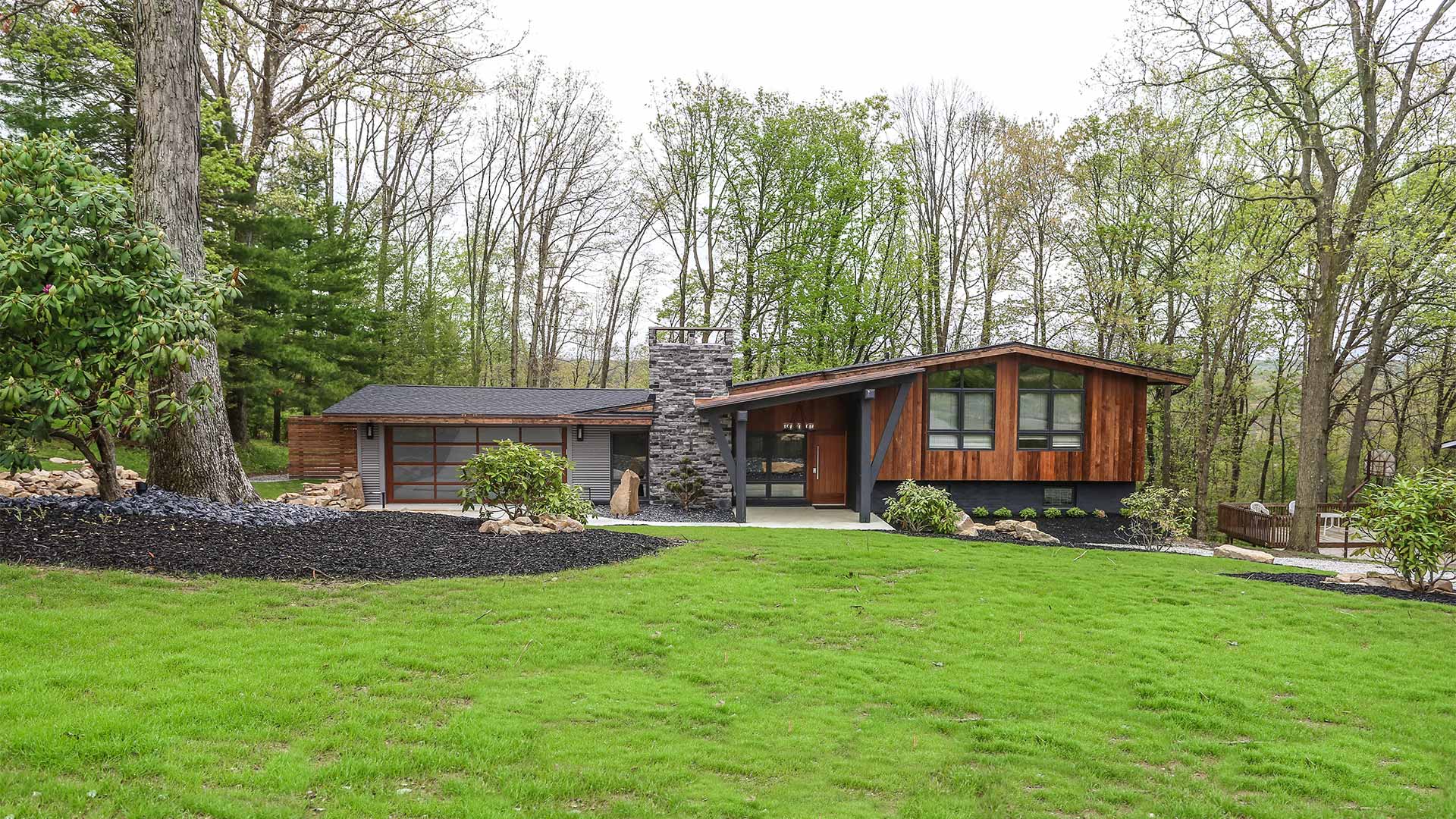 an exterior shot of the greystone home. The house has a dark wood exterior. There is nice landscaping along the front of the house and there are tall trees in the background.