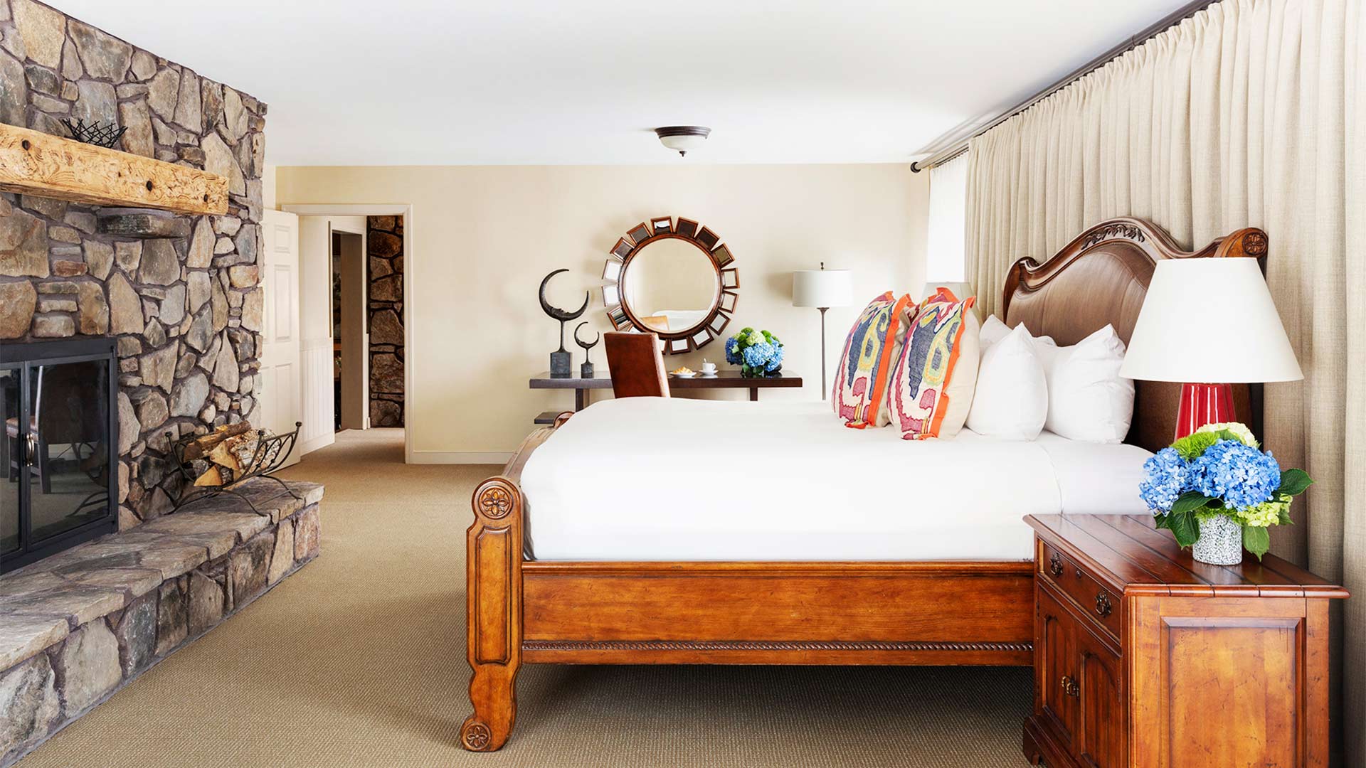 interior shot of a bedroom with a king bed with white linen and colorful throw pillows. There is a stone fireplace across from the bed. There is a vanity on the other side of the room.