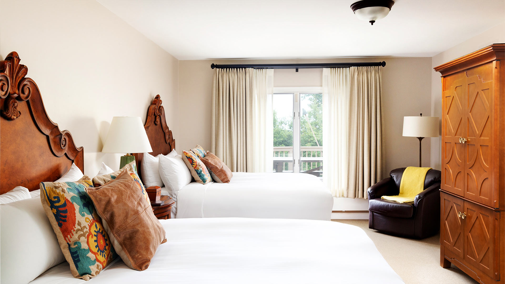 a bedroom with two queen beds. Both beds have white bedding and colorful throw pillows and intricate wooden headboards. There is a armoir and small sitting area on the other side of the room