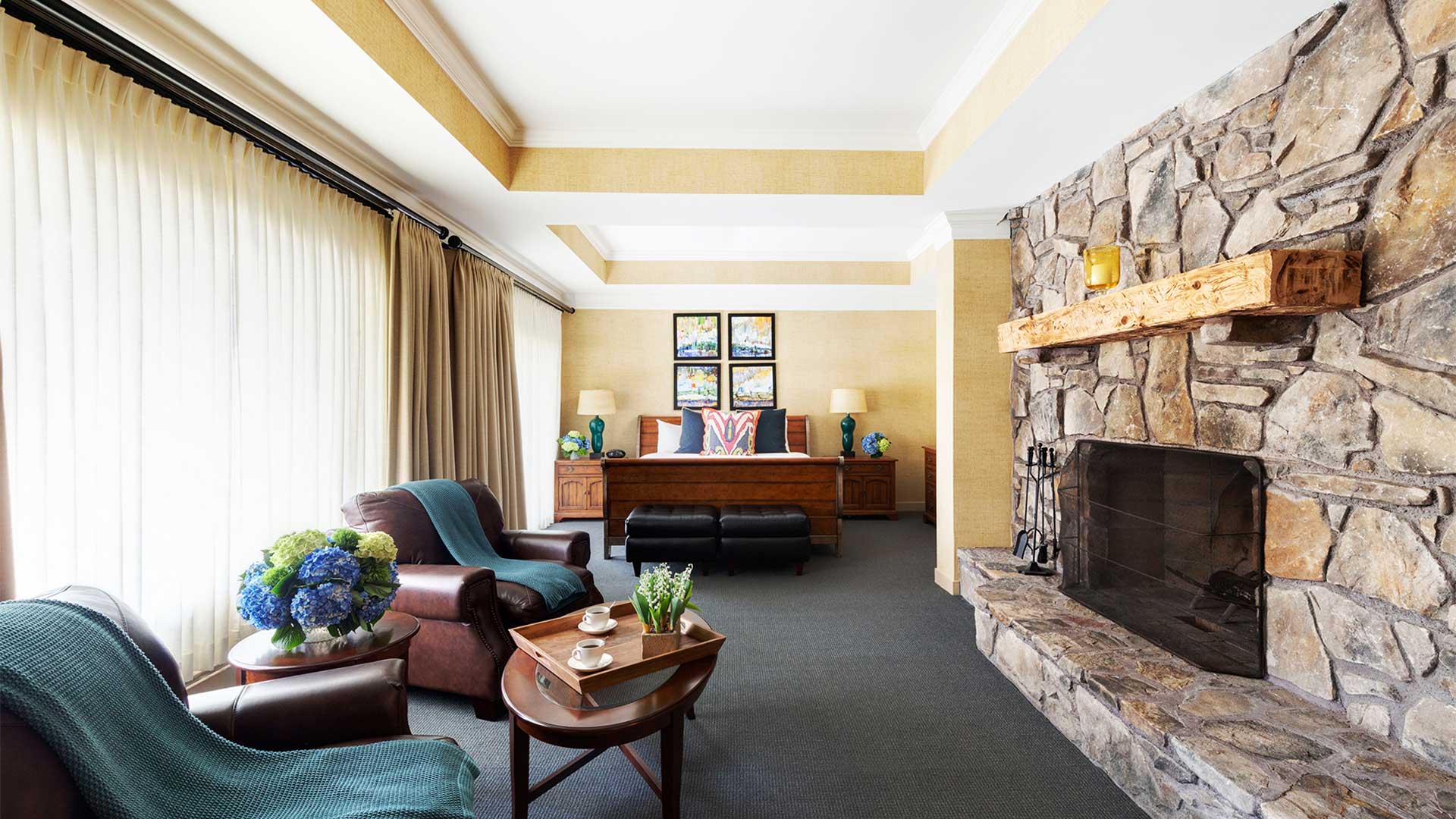 interior shot of a bedroom. There are brown leather chairs across from a large stone fireplace. There is a bed on the other side of the room with tables on either side of it.