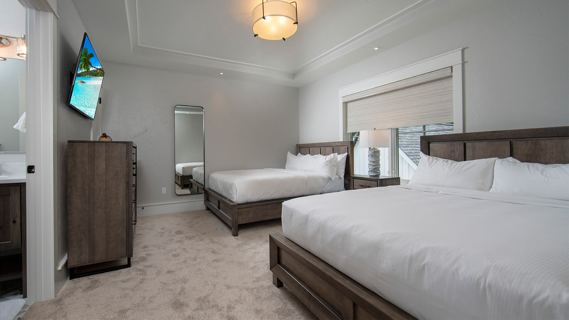 interior shot of a bedroom with two queen beds. Each bed has all white linens and wooden headboard. There is a dresser with a tv mounted overhead across from the beds. A door is open revealing a bathroom