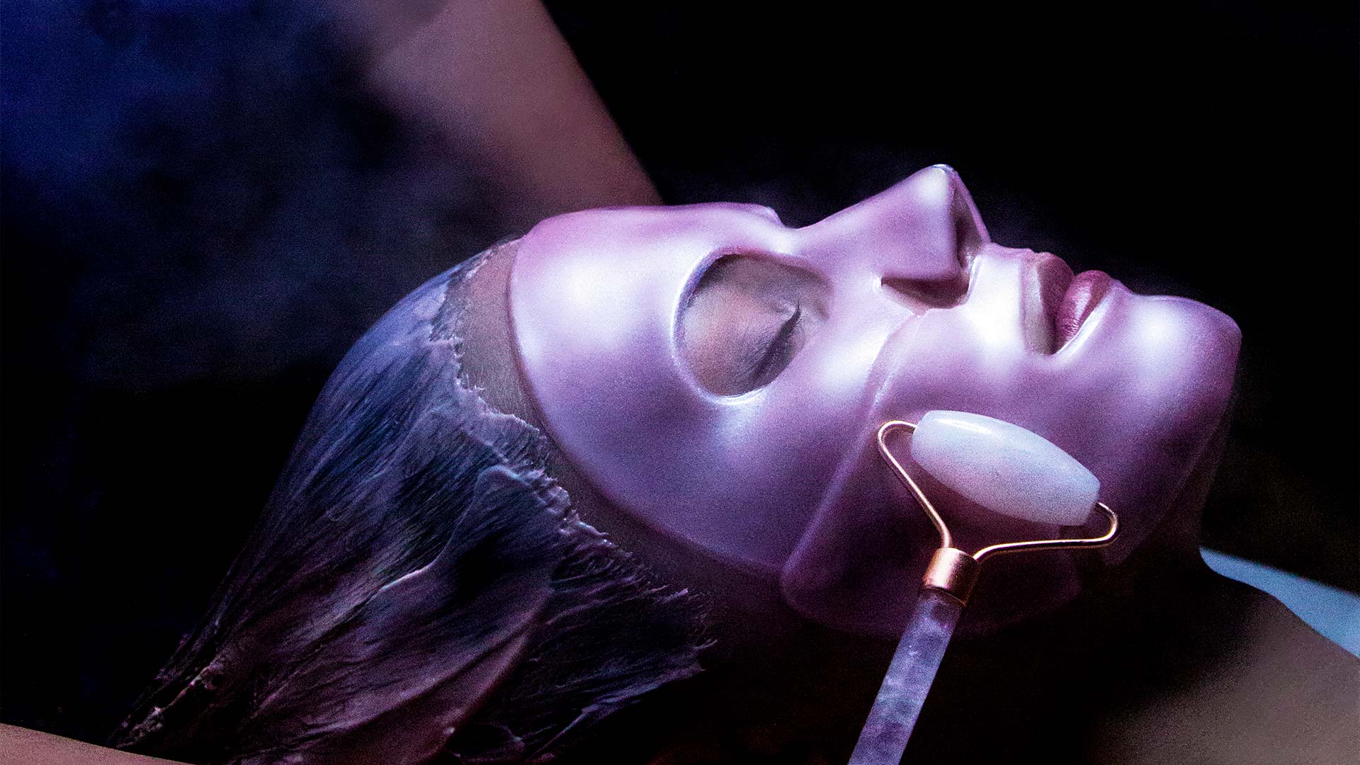 close up of a woman getting a spa treatment done. She has a pink mask on her face and a jade roller is moving across her face.