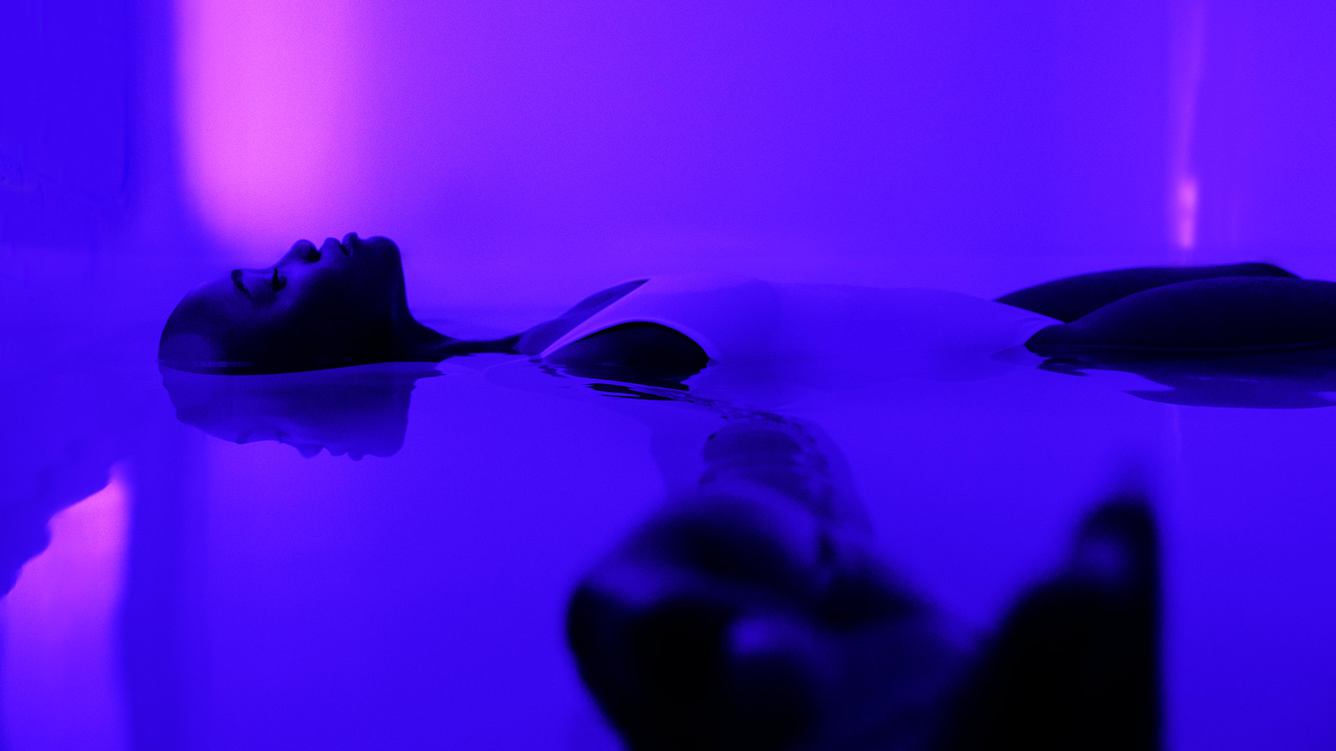 a woman laying face up in a pool of water. There lighting is a calming purple-blue