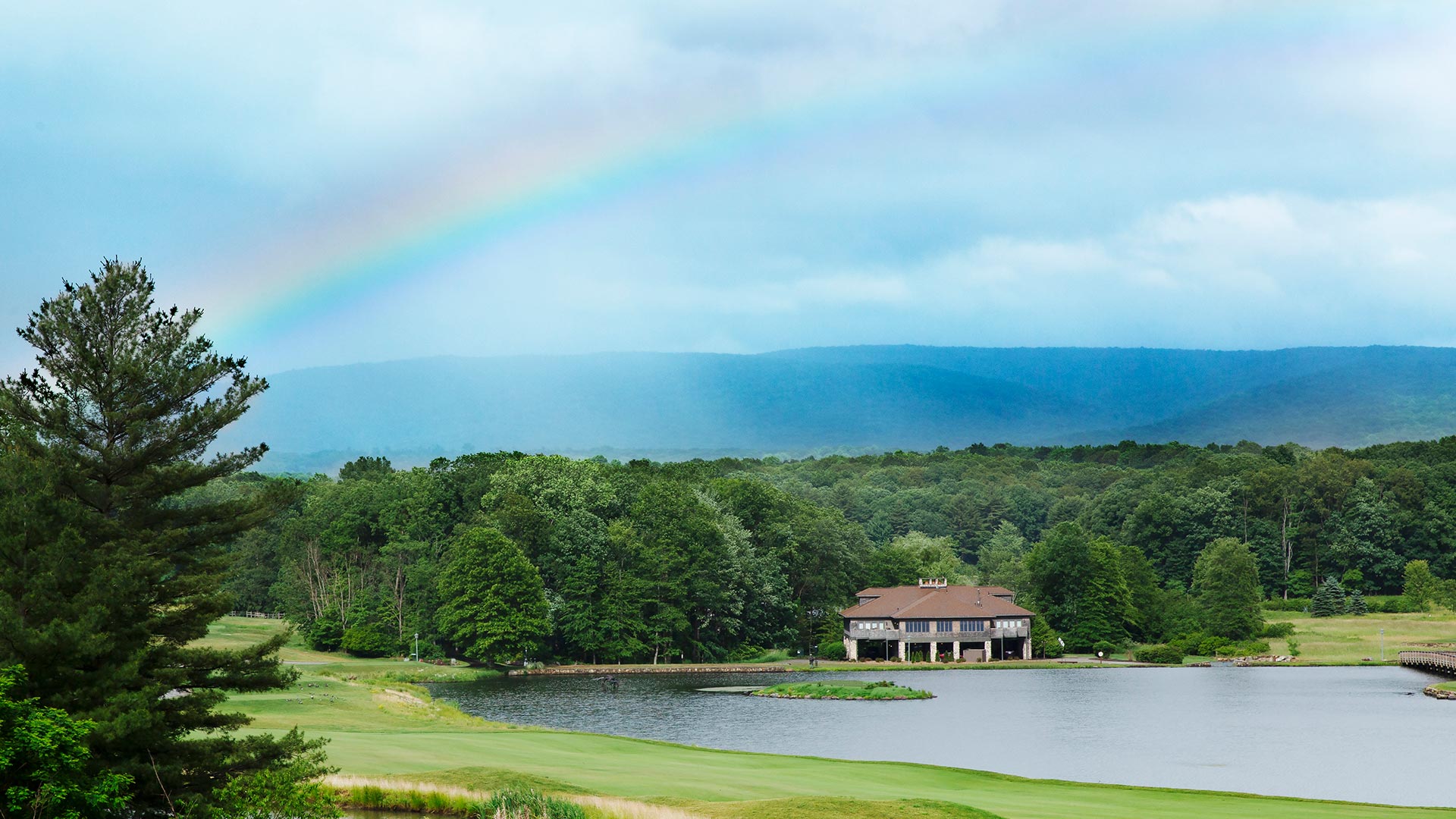 a house is perched on the lake's edge and is surrounded by forests and the golf course. There is a rainbow across the sky