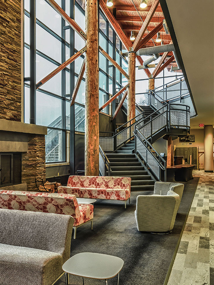 area with couches, wooden beams, a staircase, and a stone fireplace with big picture windows