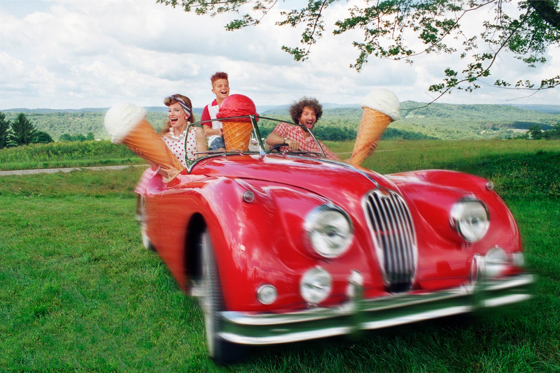 A family in a vintage red car in a field holding up giant ice cream cones