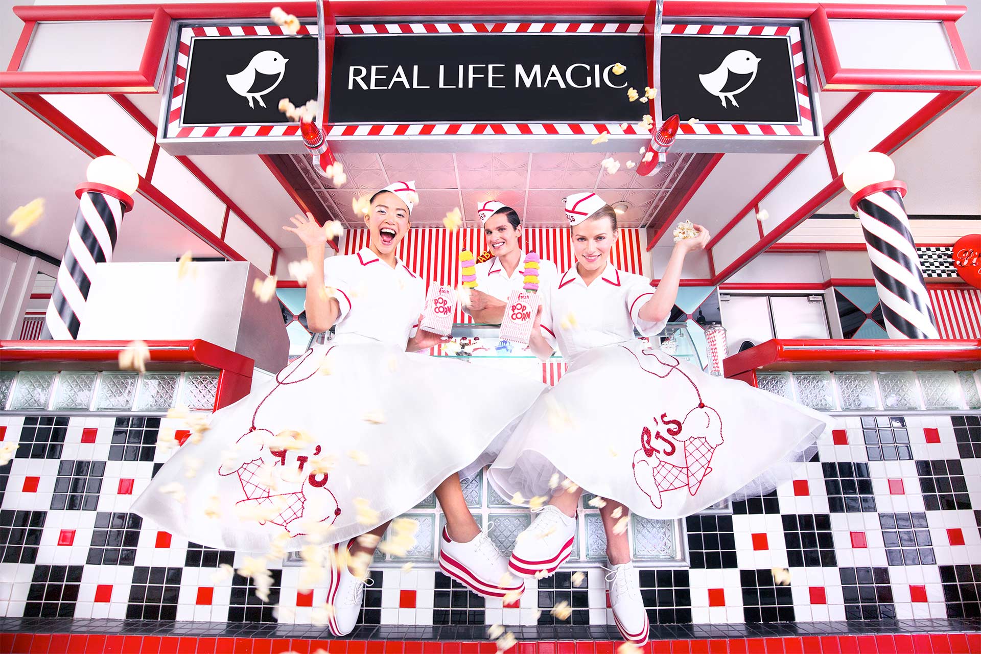 Three women dressed in 60's style uniforms in front of an ice cream shop with a sign that says "Real Life Magic"
