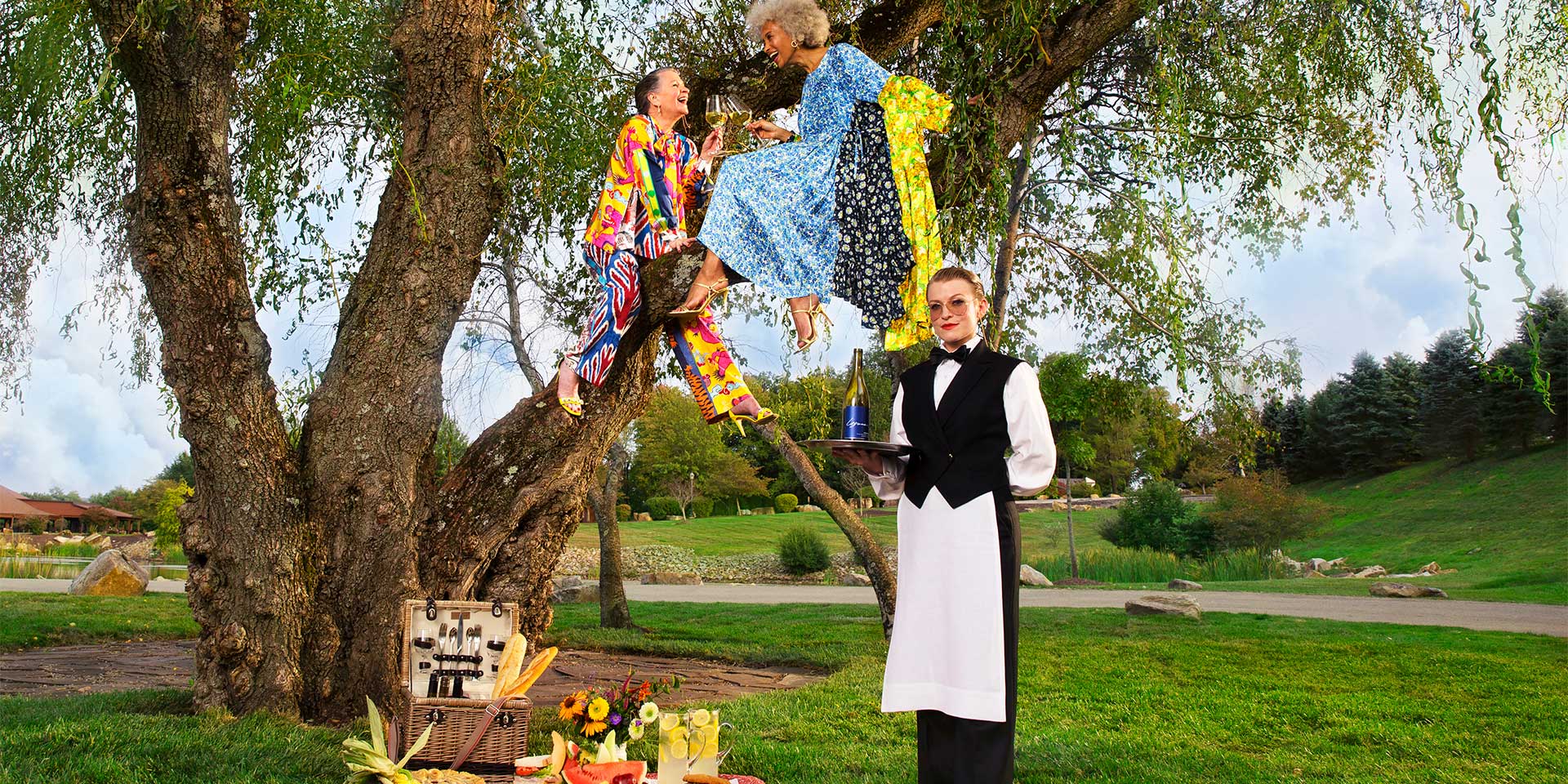 Two older women sit in a tree and enjoy a picnic on the ground catered to them by a woman standing in front of them with champagne in hand.