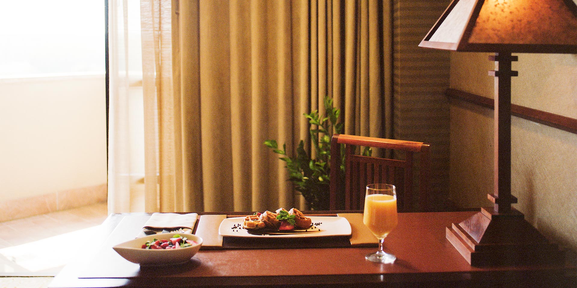 Waffles, orange juice, and a bowl of fruit is placed on a brown wooden table in a well light room.