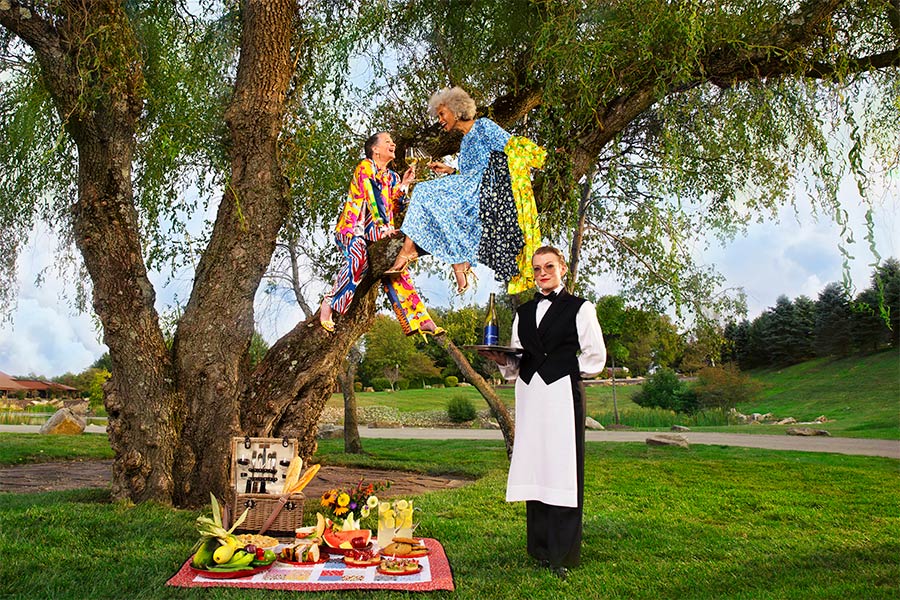 Two older women sit in a tree and enjoy a picnic on the ground catered to them by a woman standing in front