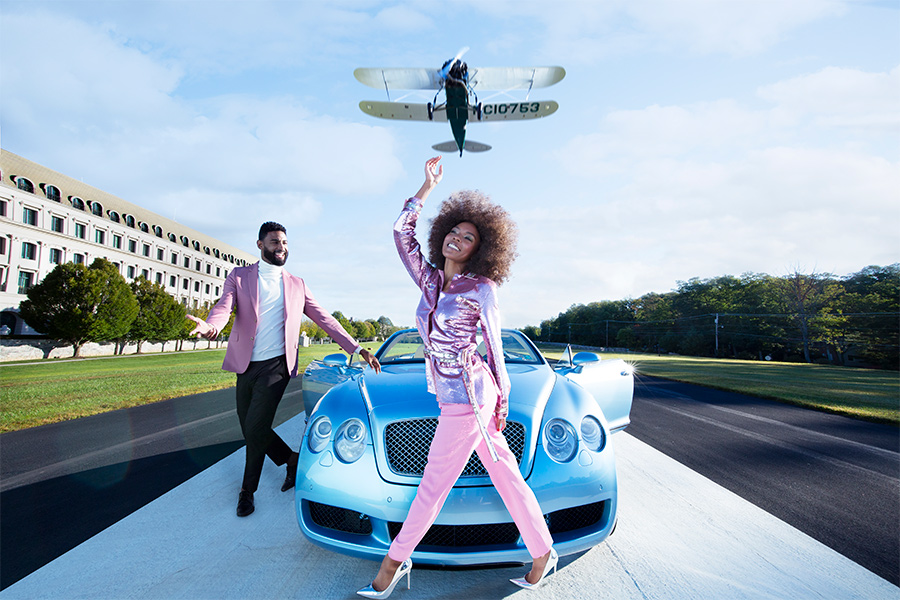 A woman wearing pink standing in front of a blue car with a man wearing pink next to the car and a plane flying above