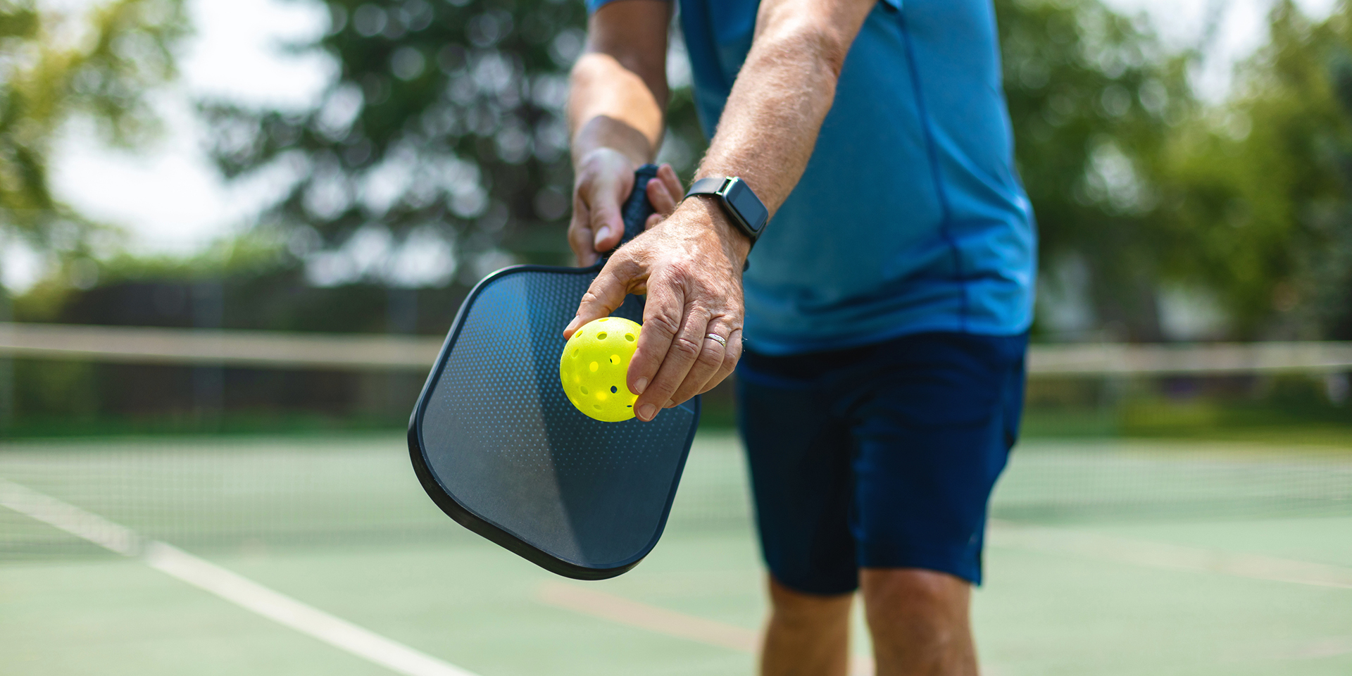 Mature adult male in a blue shirt and shorts preparing to serve a yellow pickleball with a blue paddle