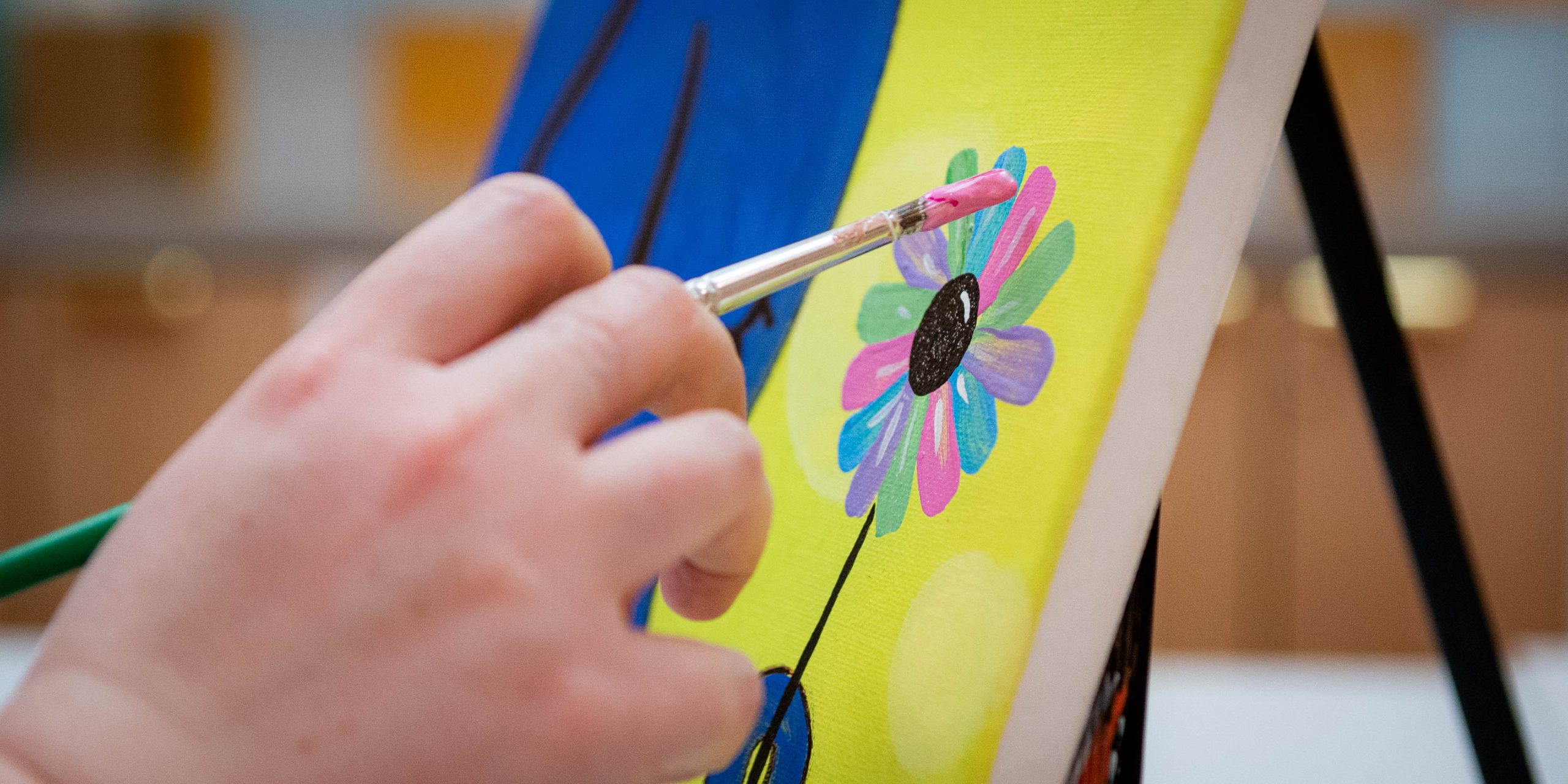 A hand holding a paint brush, painting a multi-colored flower on canvas.