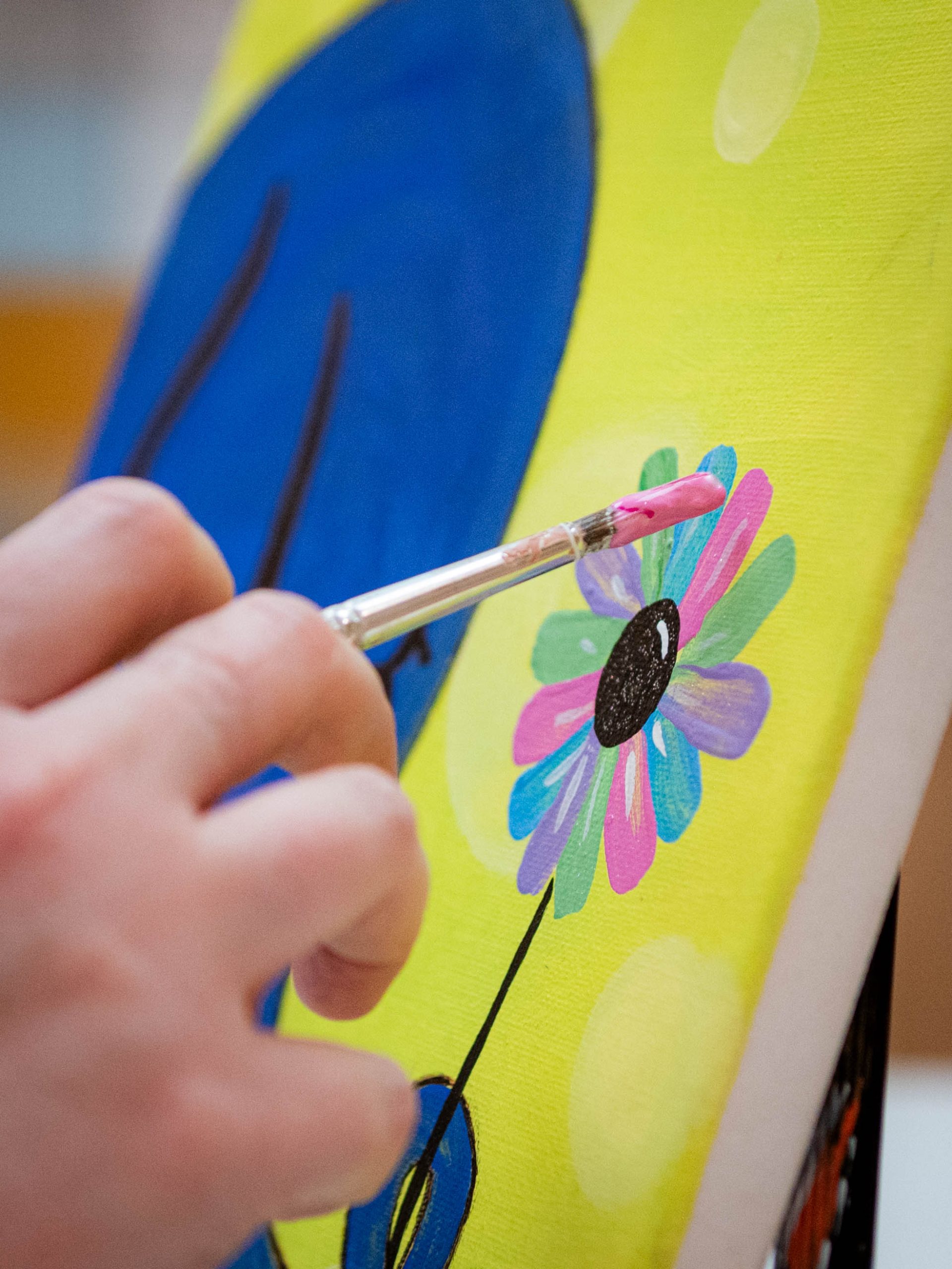 A hand holding a paint brush, painting a multi-colored flower on canvas.