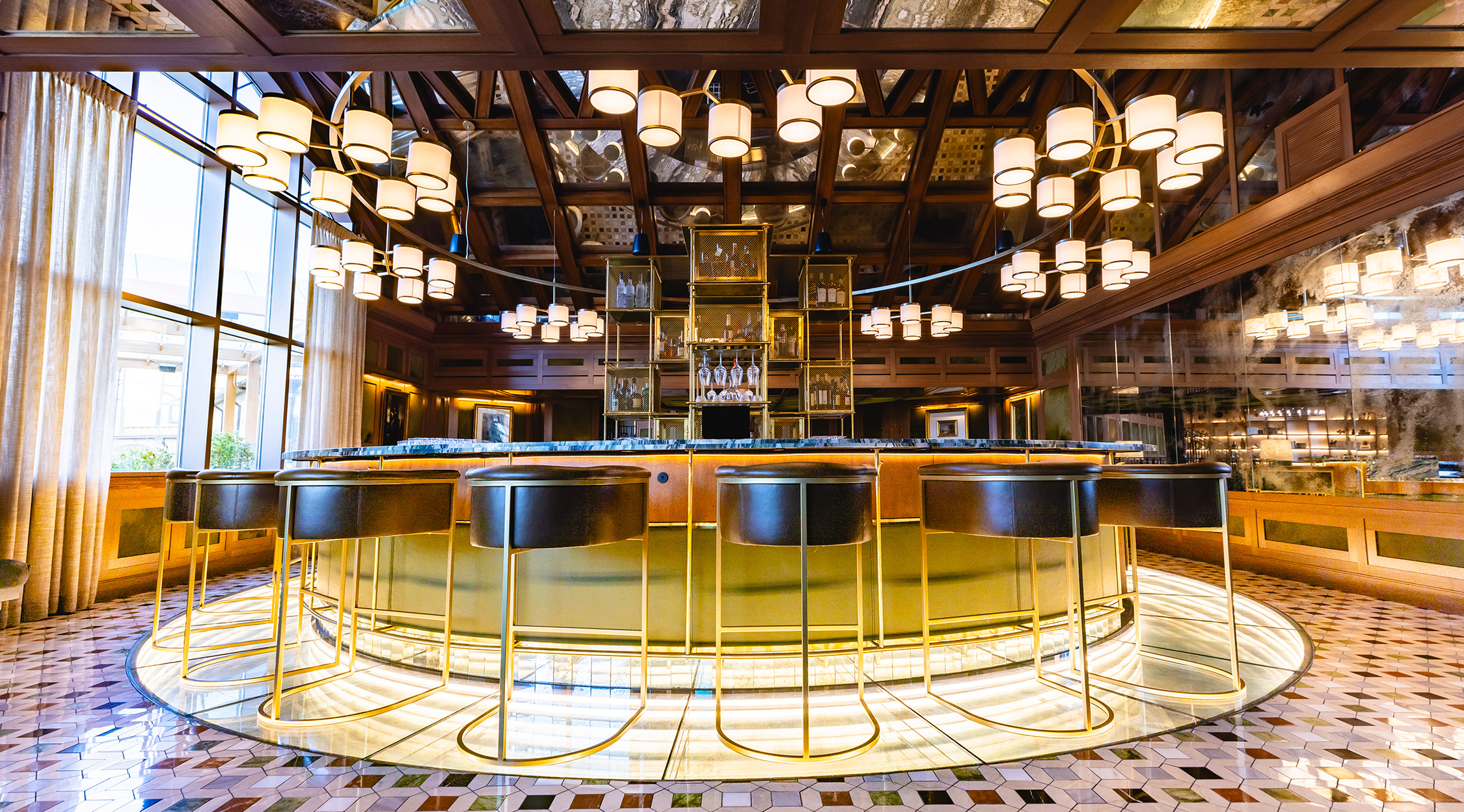The Circle Bar at The Grand Lodge at Nemacolin features a circular bar surrounded by bar stools, mosiac tile and mirrored flooring, and numerous overhead chandeliers in a modern space that houses a mid-century feel