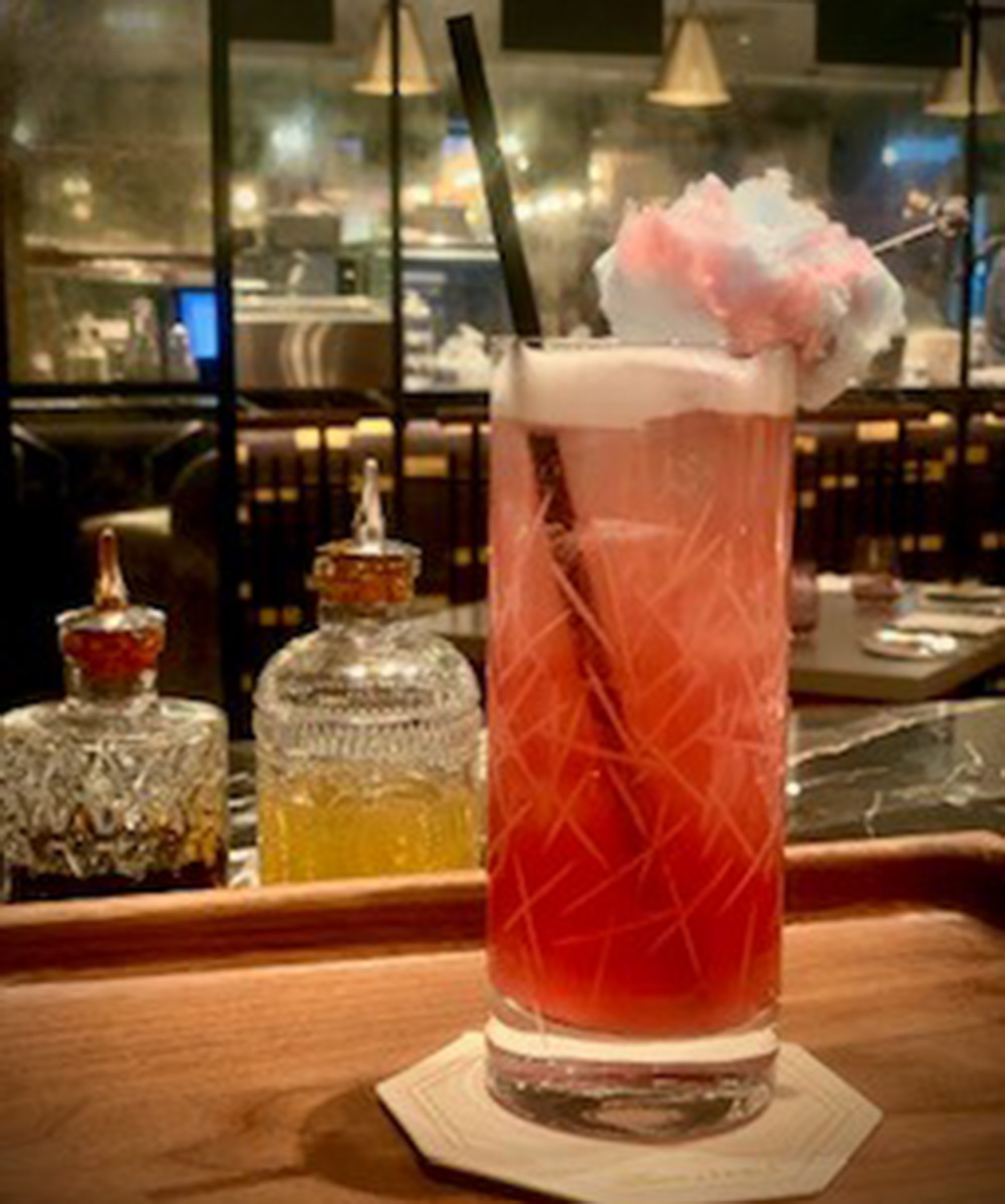 Reddish-pink mocktail in a tall glass with a straw and a garnish of a puff of cotton candy