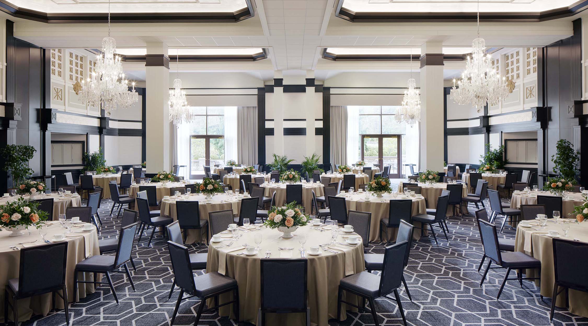 A ballroom with crystal chandeliers, deep-toned wood, light carpeting, and high ceilings decorated with round tables in the Grand Ballroom at Nemacolin resort