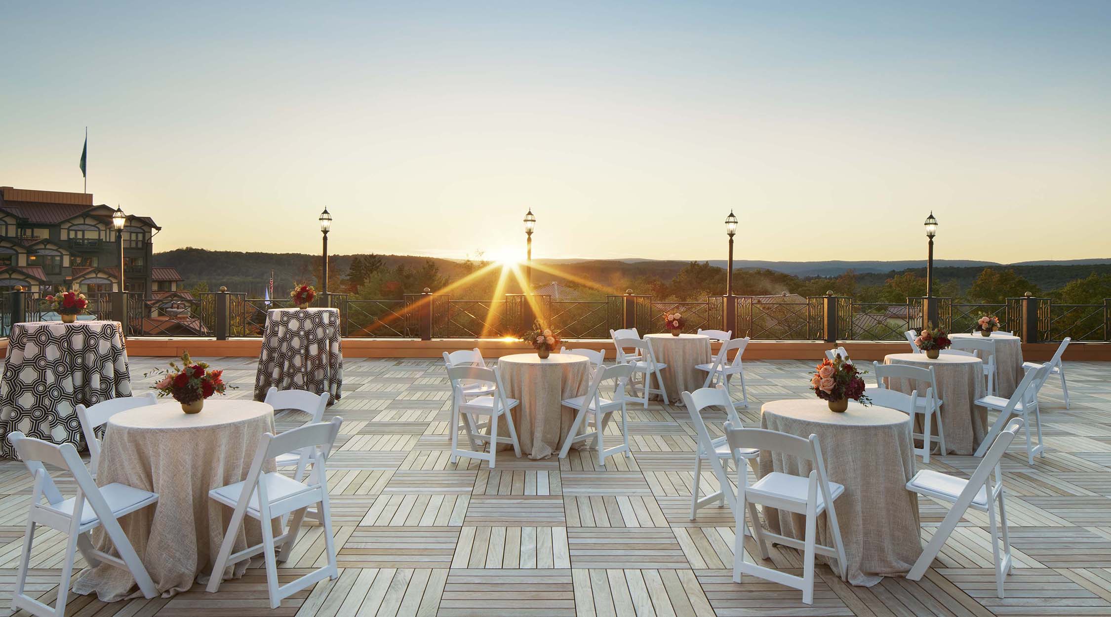 Small round tables and white chairs on a rooftop deck of parquet wood, overlooking a hotel, mountains, and sunset rays, on the Plaza Deck located at Nemacolin resort