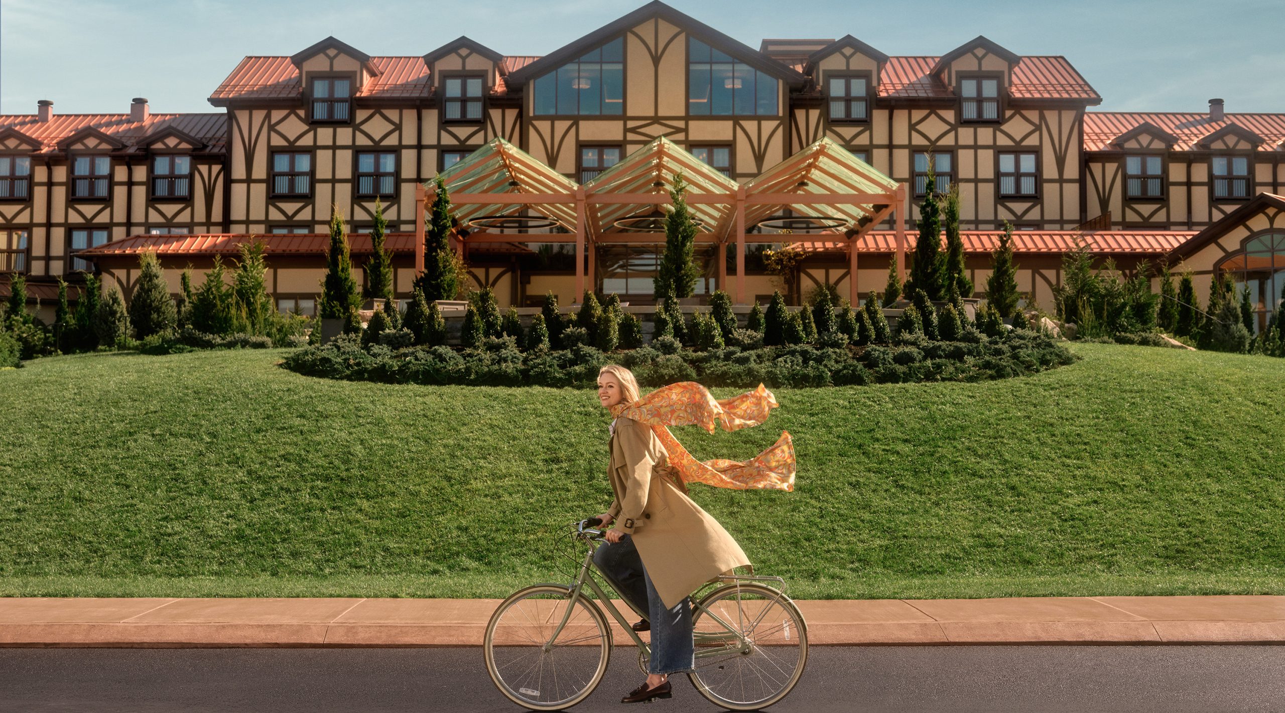 A woman wearing a beige coat and an orange scarf riding a bike in front of The Grand Lodge at Nemacolin resort