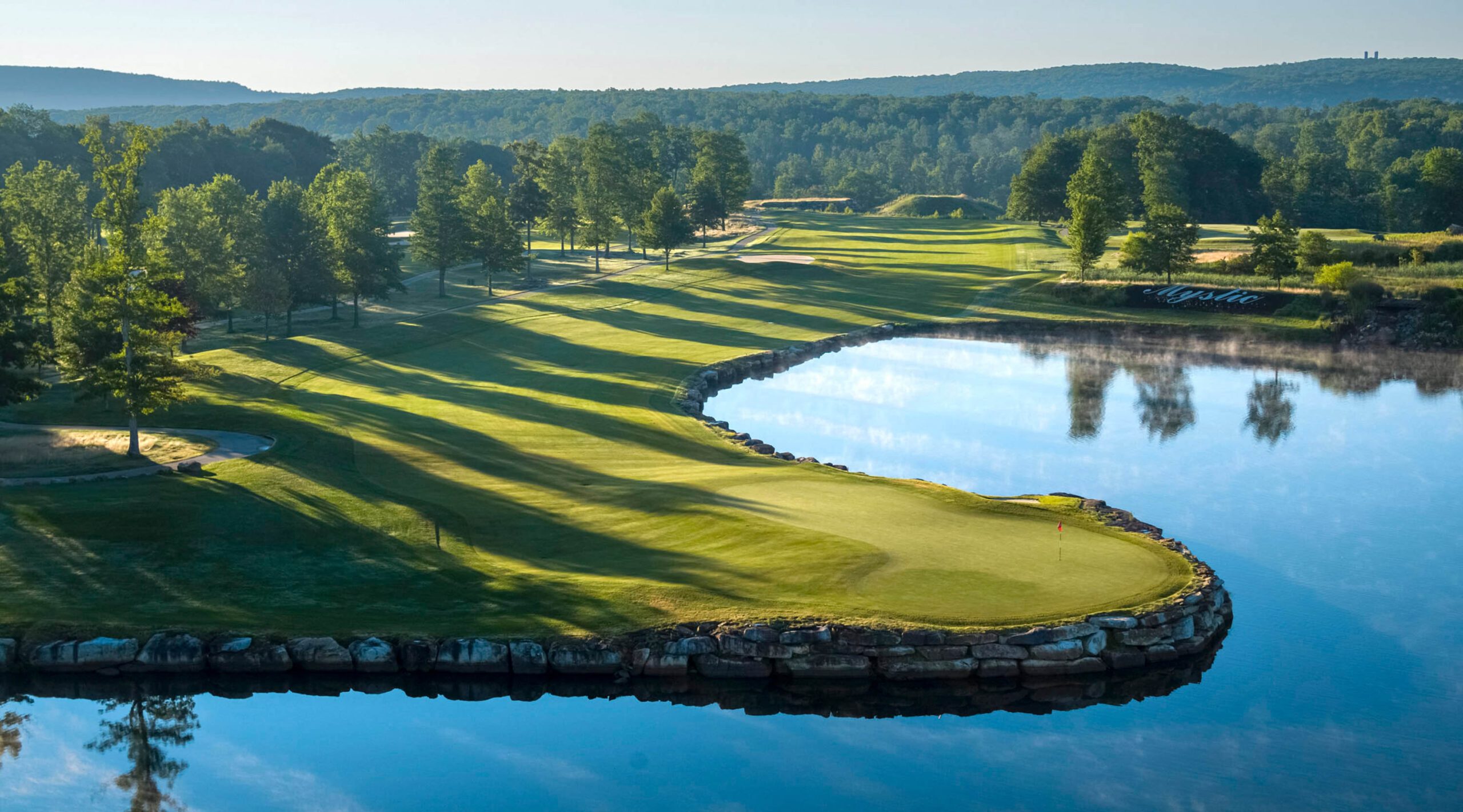 A landscape of a fairway, green, lake, and mountains on the Mystic Rock golf course at Nemacolin resort