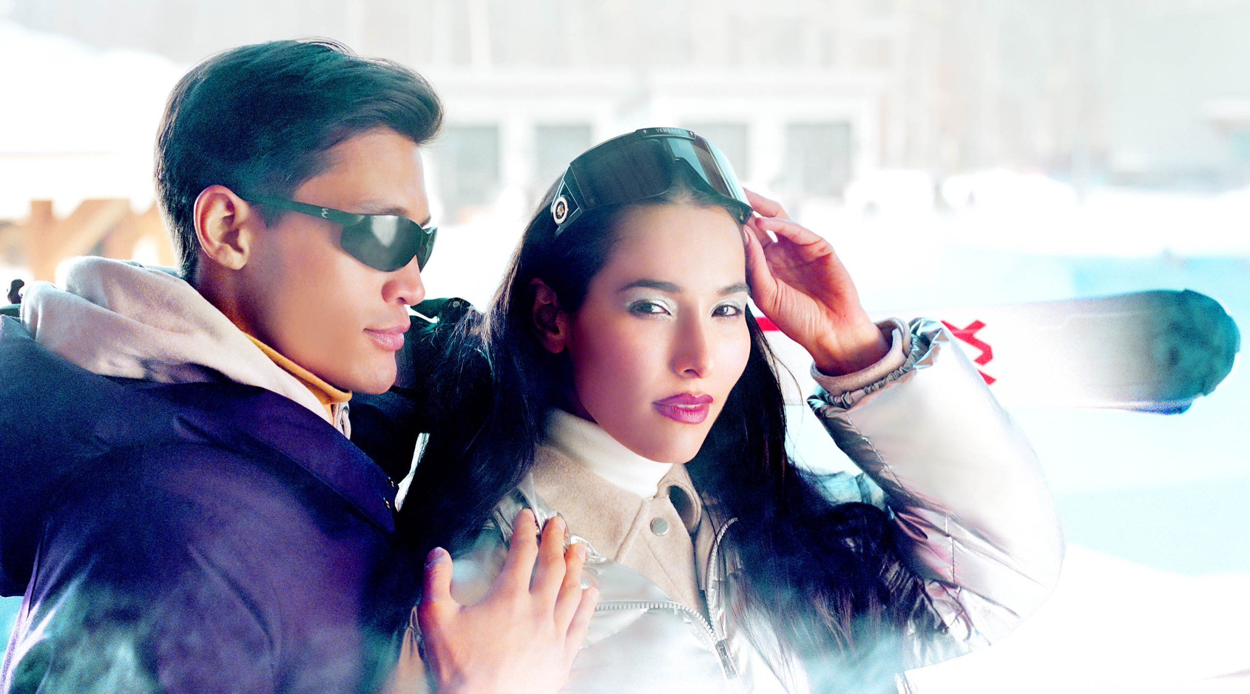 A man with sunglasses and a purple winter coat with his hand on the shoulder of a woman with sunglasses on her head who is wearing a white winter coat