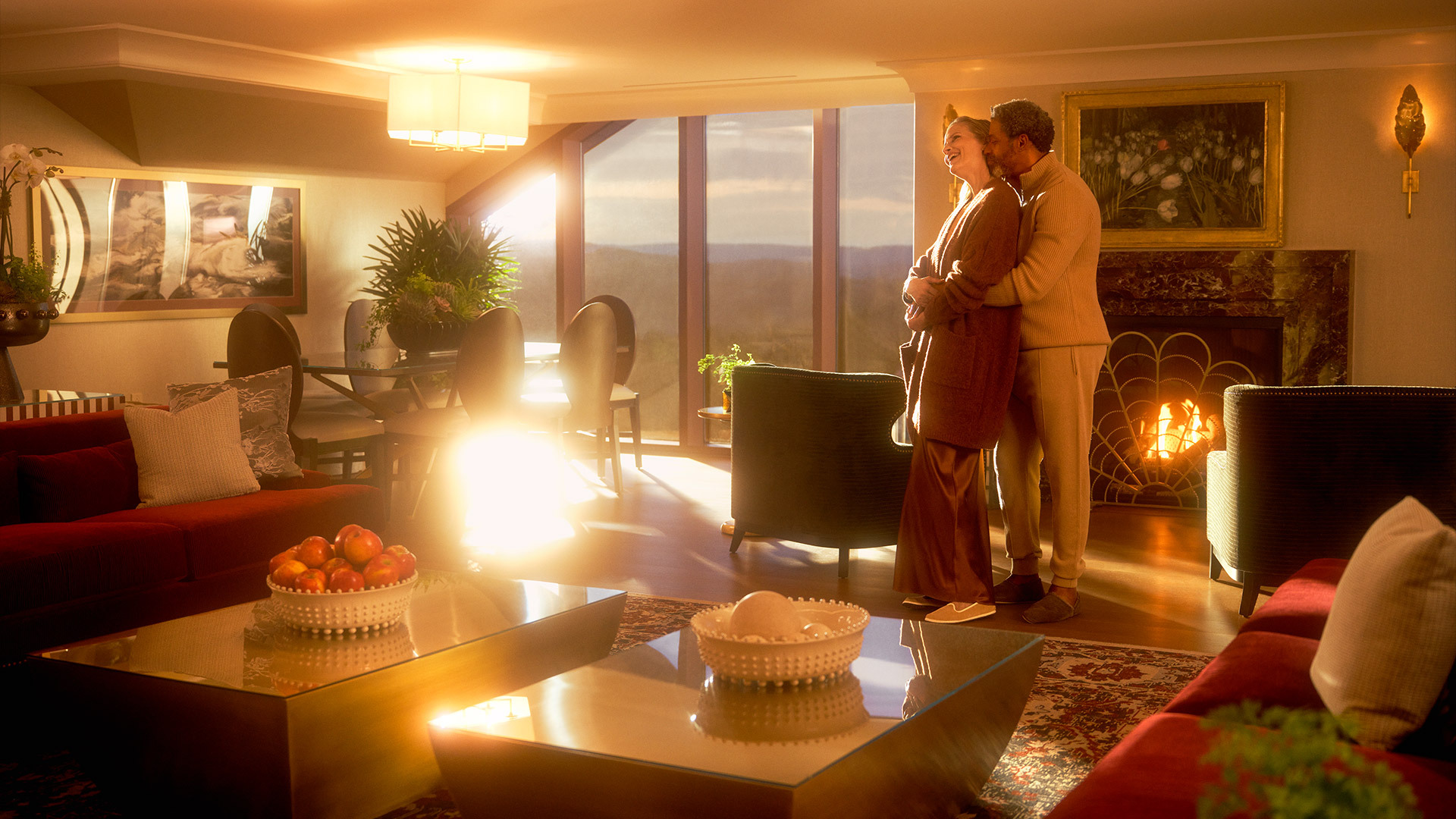 A man embracing a woman from behind standing in a hotel room in front of a fireplace near a floor-to-ceiling window