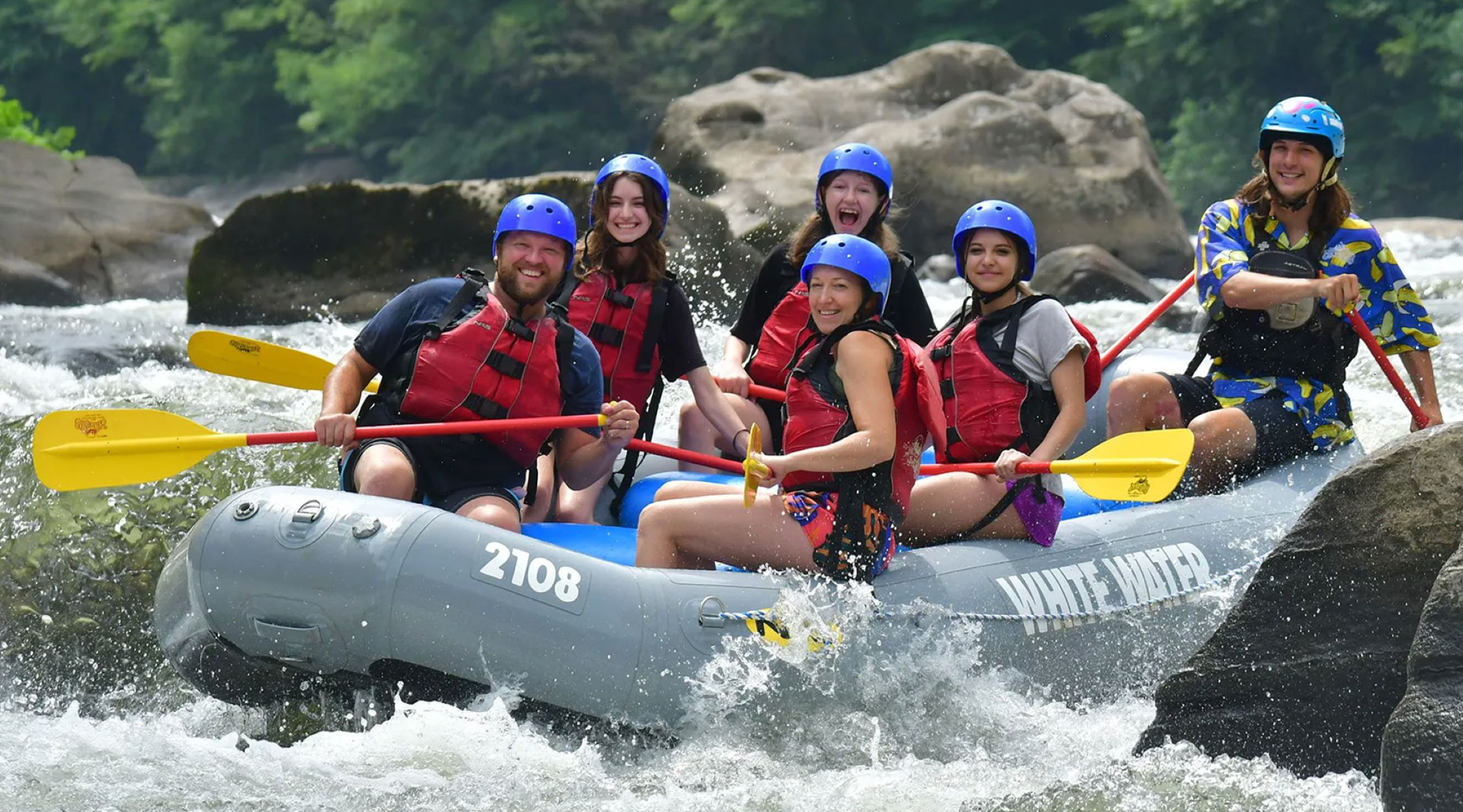 Four women and two male guides wearing blue helmets and red flotation vests white water rafting in an inflatable grey raft on the Yough River near Nemacolin resort