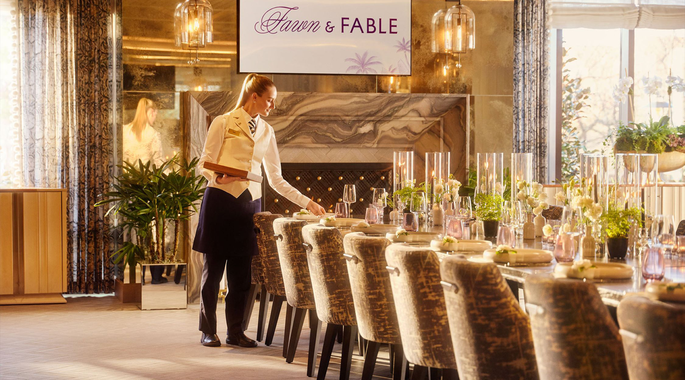 A female restaurant server dressed in a cream-colored vest and shirt with black pants and a black apron setting a long dining table which will seat more than a dozen people that is located in front of a fireplace in the Fawn & Fable restaurant in The Grand Lodge at Nemacolin