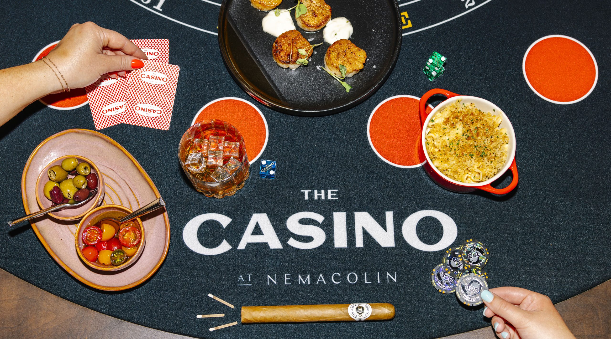 Photo of a blackjack table at The Casino Nemacolin that also two womens' hands, playing cards, casino chips, dice, a cigar with matches, and an assortment of food, including scallops, olives, tomatoes, and a casserole.