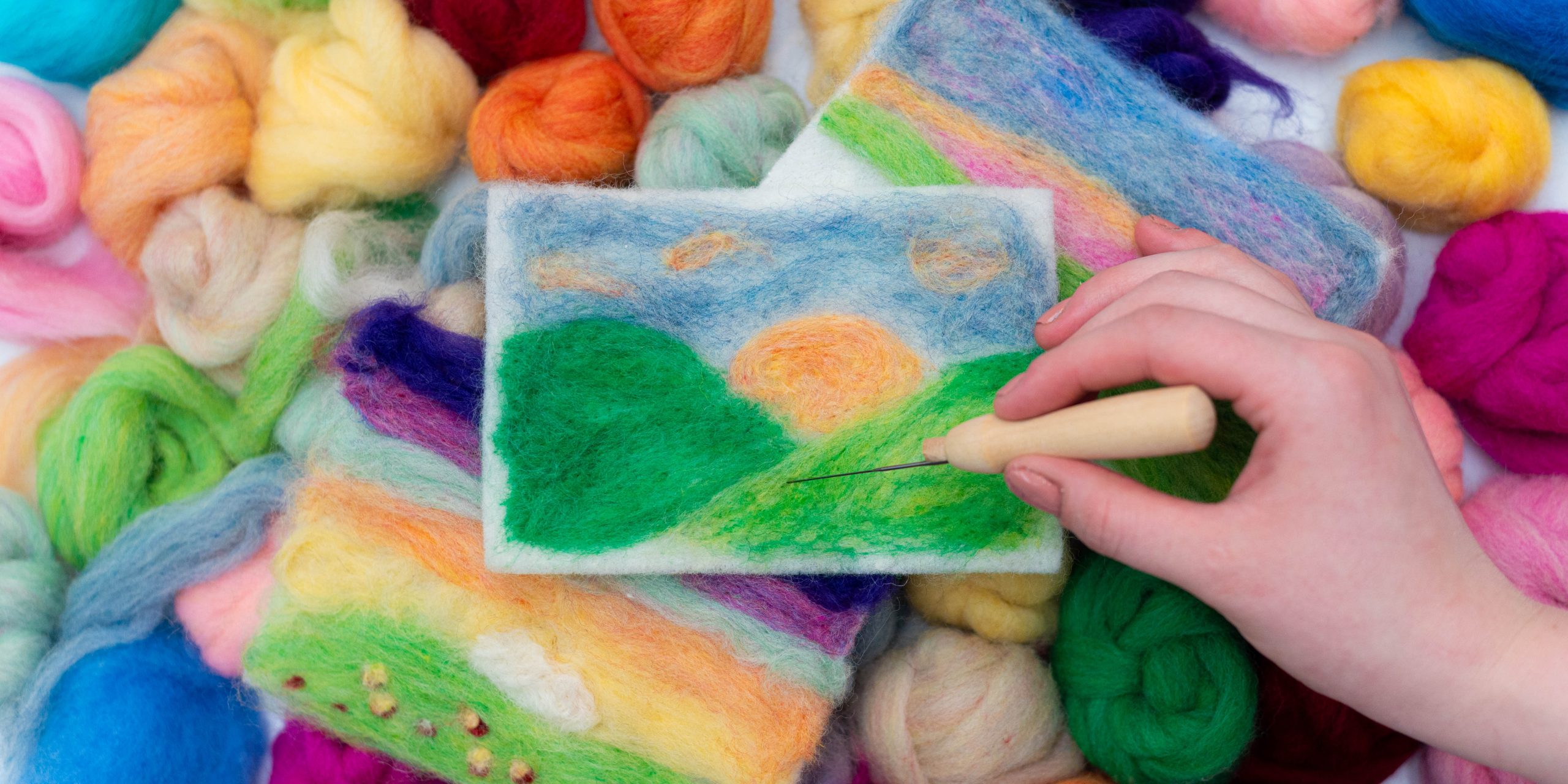 A hand reaches in from the right side with a tool to create a landscape image with various colors of wool.