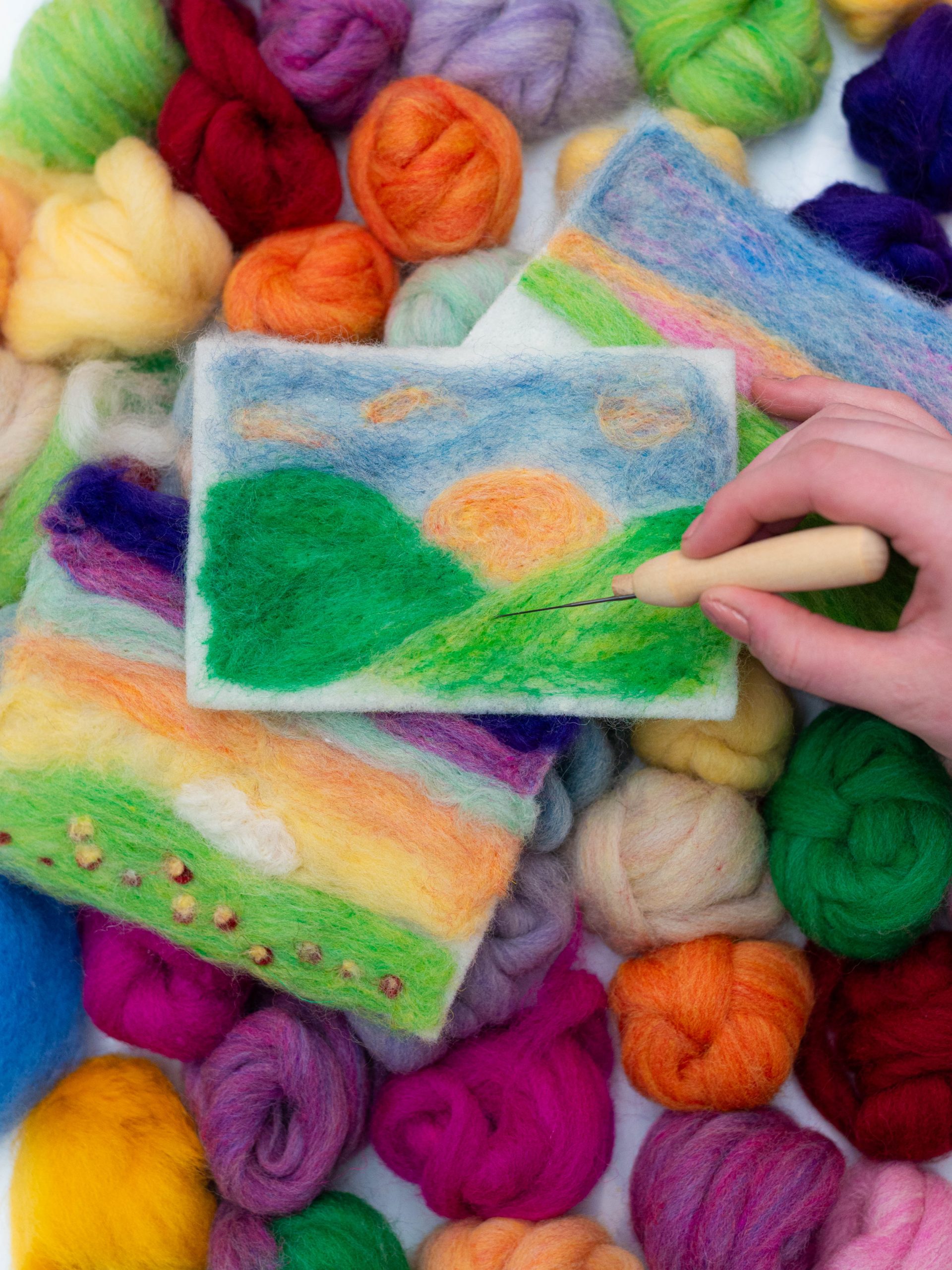 A hand reaches in from the right side with a tool to create a landscape image with various colors of wool.