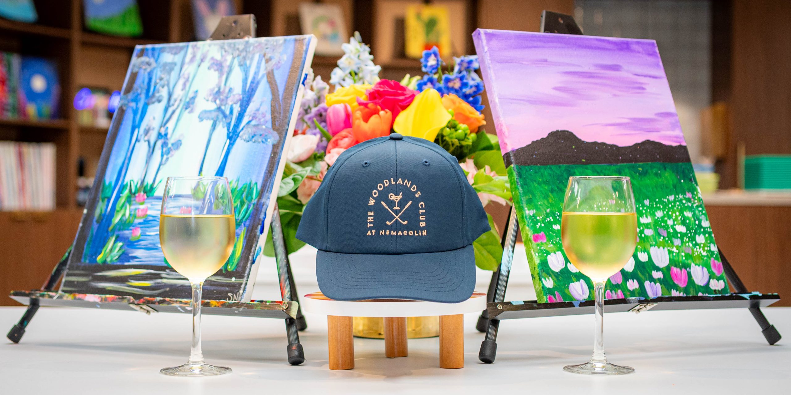 A blue hat with the logo of The Woodlands Club at Nemacolin is surrounded by two glasses of white wine, two canvas paintings, and a bouquet of flowers.