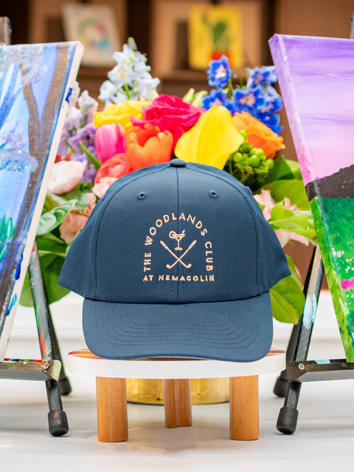 A blue hat with the logo of The Woodlands Club at Nemacolin is surrounded by two glasses of white wine, two canvas paintings, and a bouquet of flowers.
