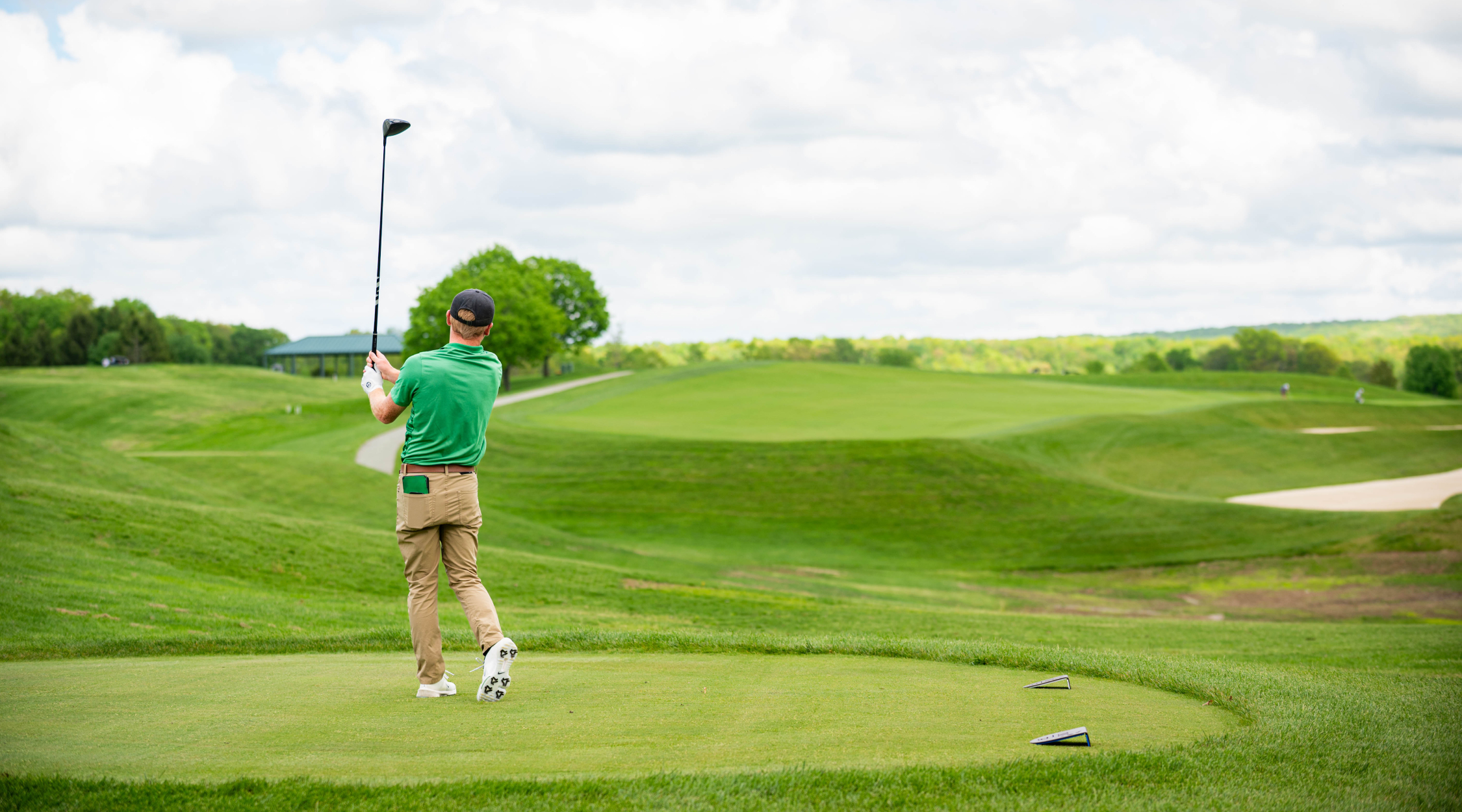 A golfer wearing a green shirt and black hat looks down the course after hitting a drive from the tee box.