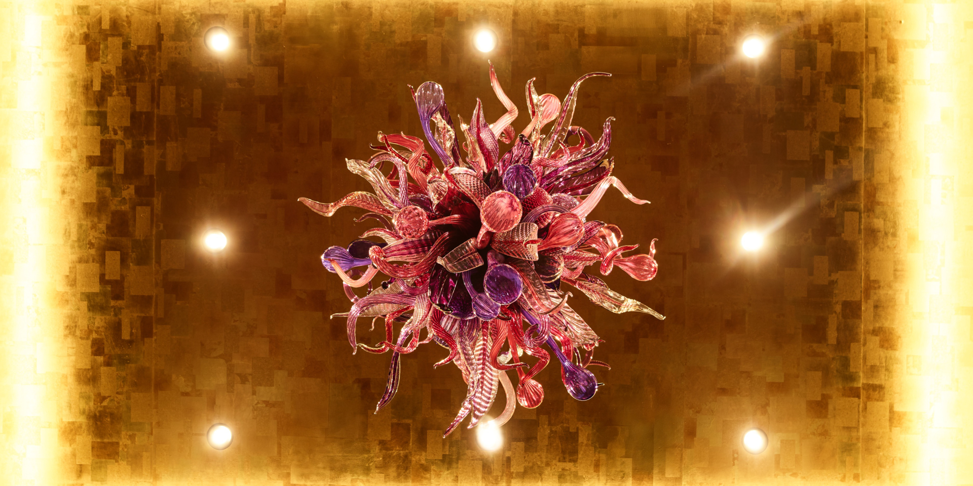 A Chihuly chandelier made up of various pink, purple, and other colors, with eight bright lights around it.