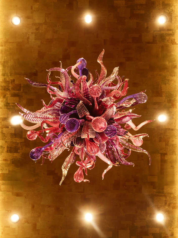 A Chihuly chandelier made up of various pink, purple, and other colors, with eight bright lights around it.