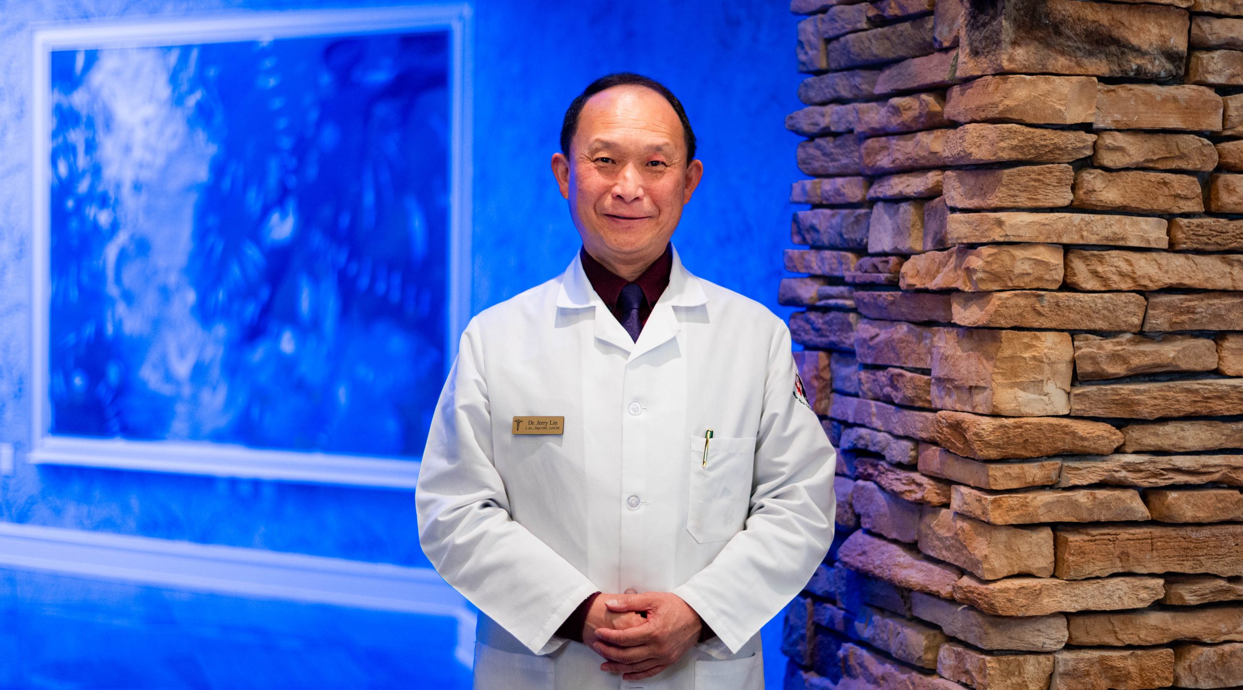 Dr. Jerry Lin smiles with his hands folded in front of him, wearing a white lab coat with a blue wall and a stone pillar behind him.