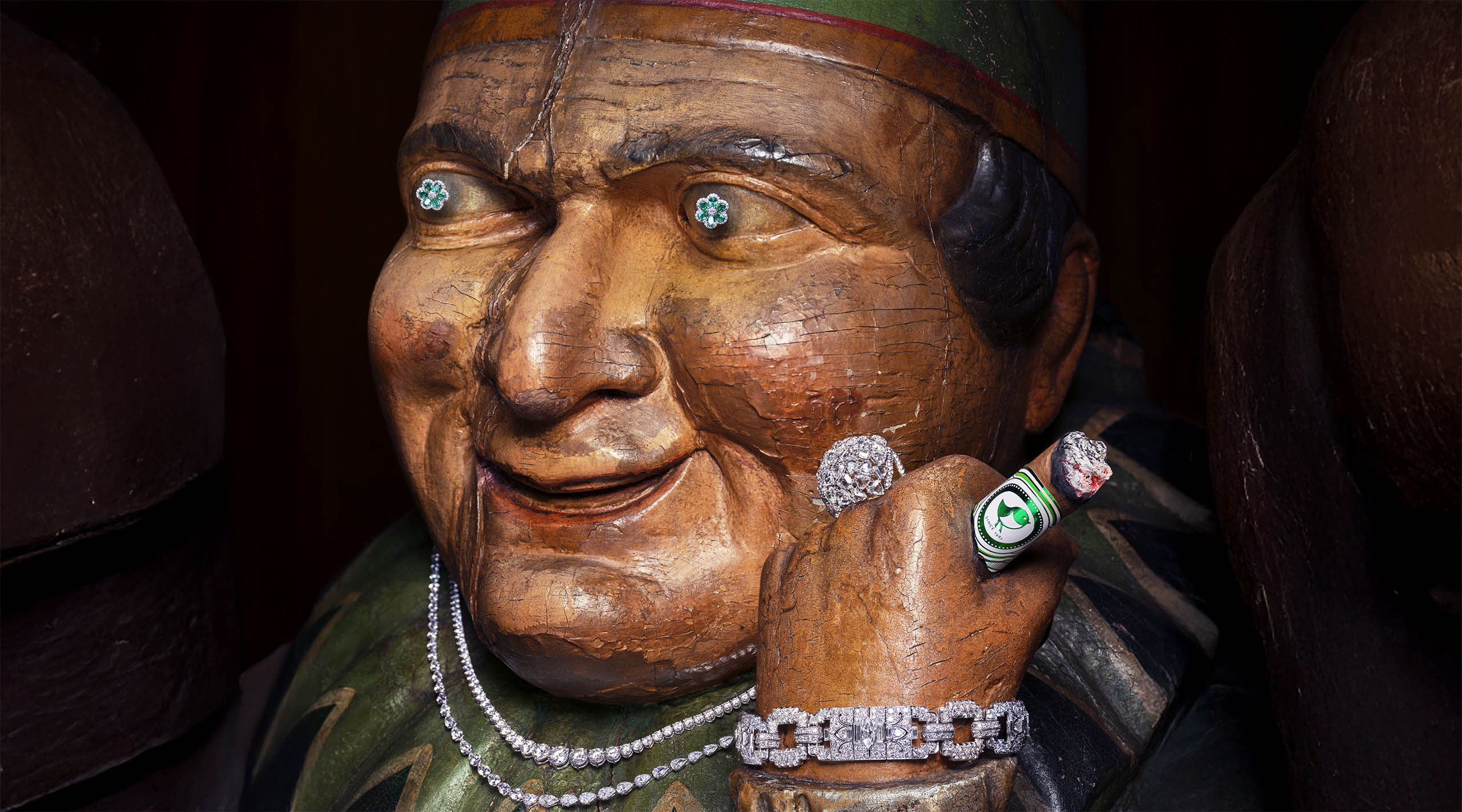 a painted wooden sculpture of a man wearing jewelry on his wrist, finger, neck, and eyes, holding a lit cigar