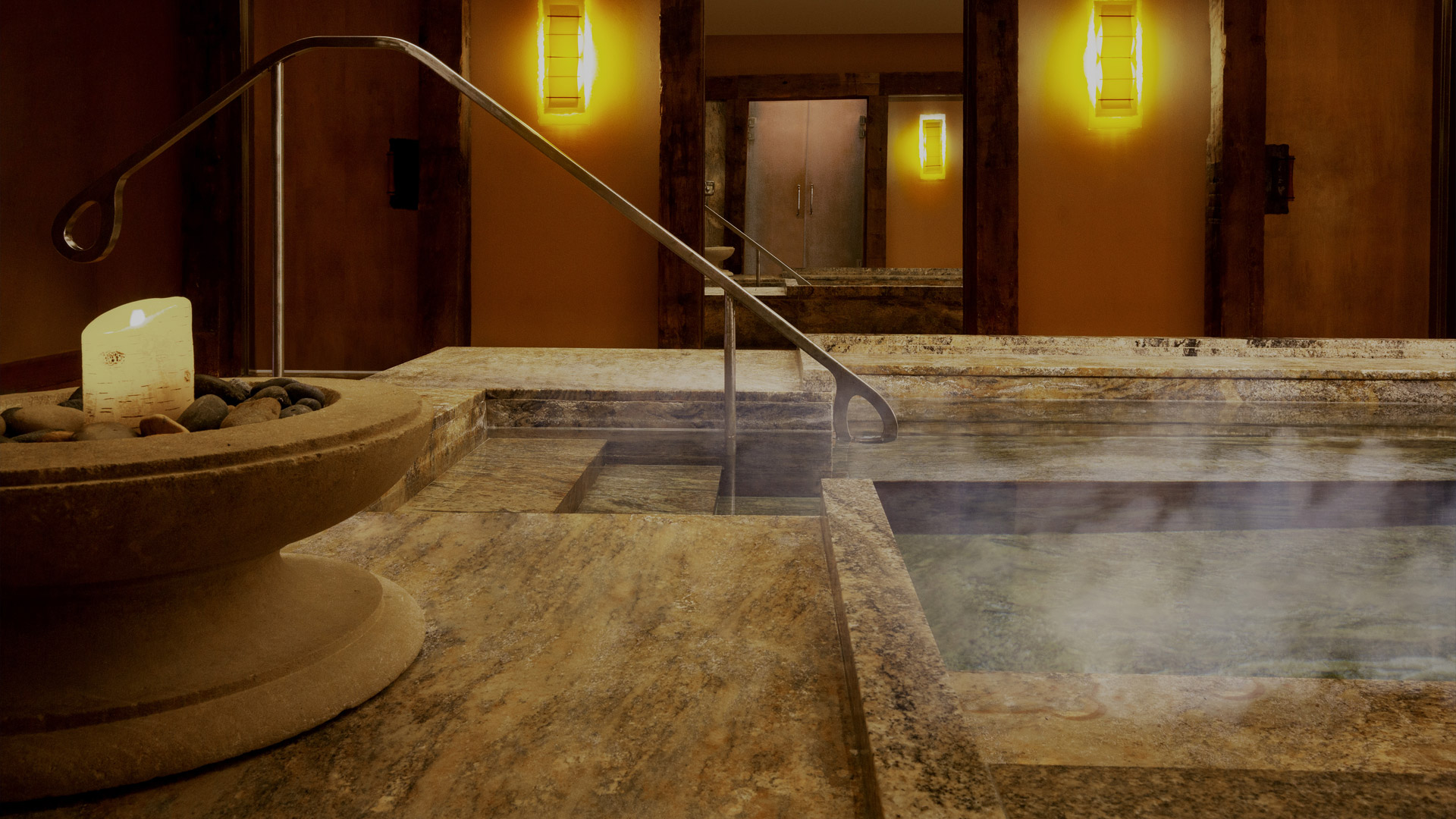 Steps entry to a brown marble pool. A candle is lit in the foreground and steam is rising from the pool.