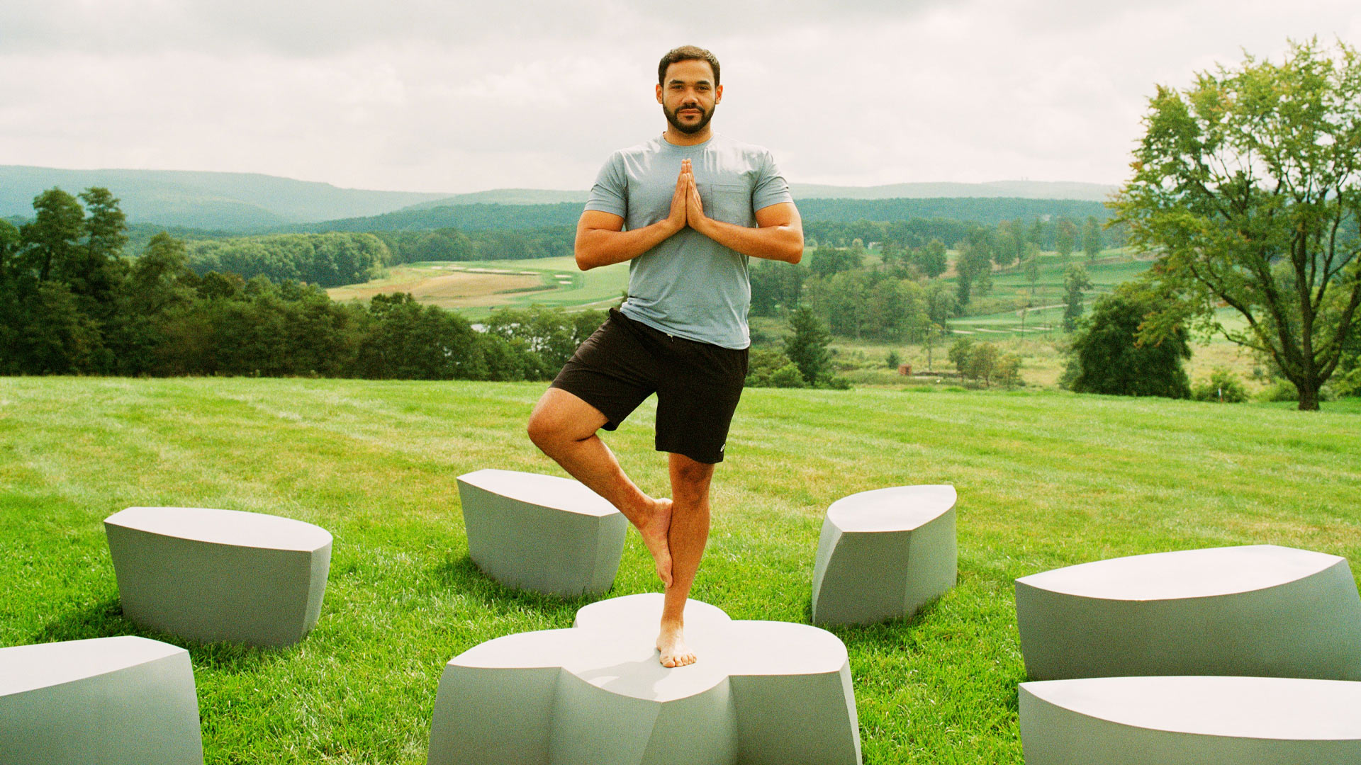 Man practicing yoga outdoors in tree pose, surrounded by modern stone seating on a lush green lawn with scenic mountain and golf course views in the background.