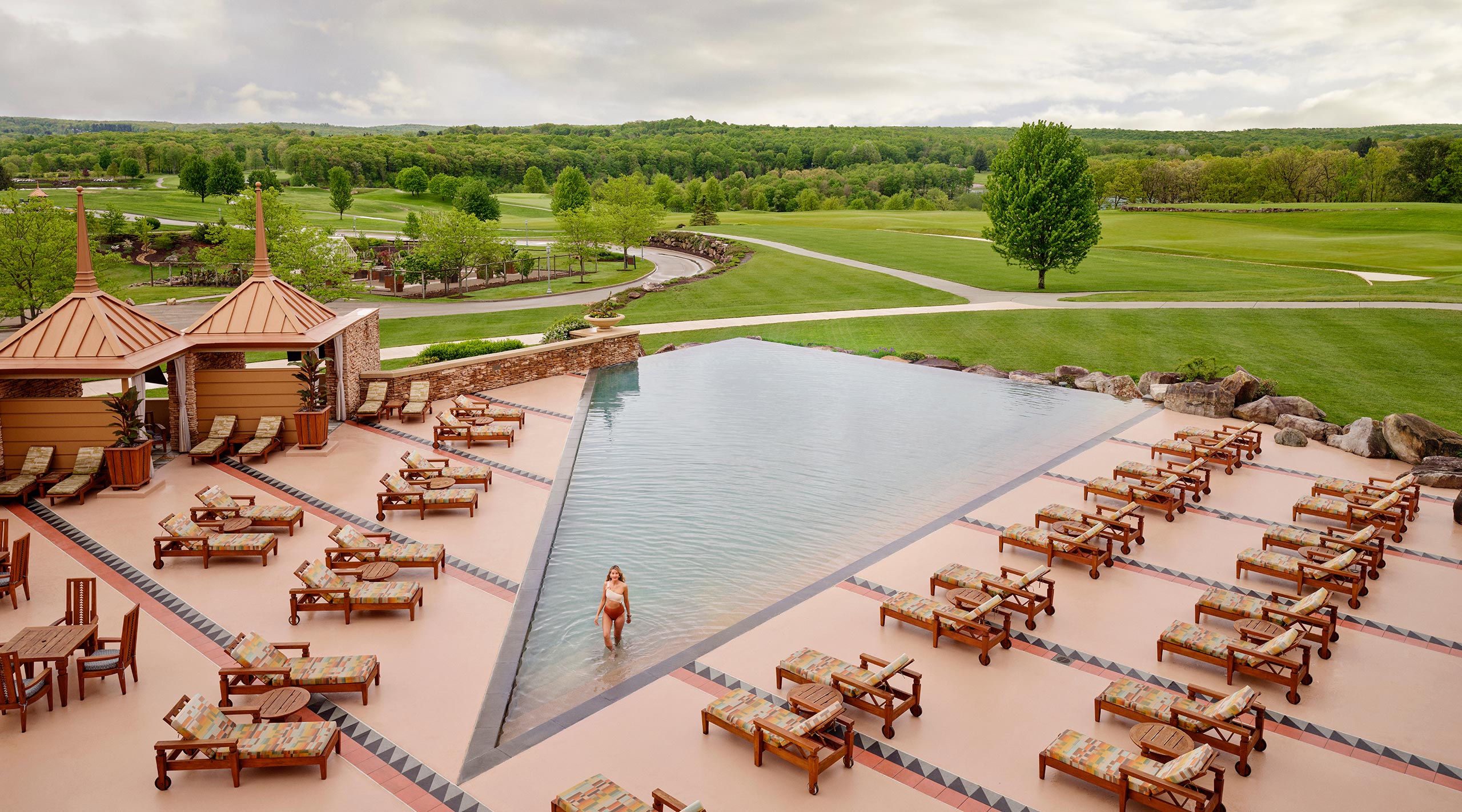 A woman walks out of a triangular-shaped swimming pool that overlooks a golf course and mountains onto the deck of the Falling Rock Infinity Pool at Nemacolin resort