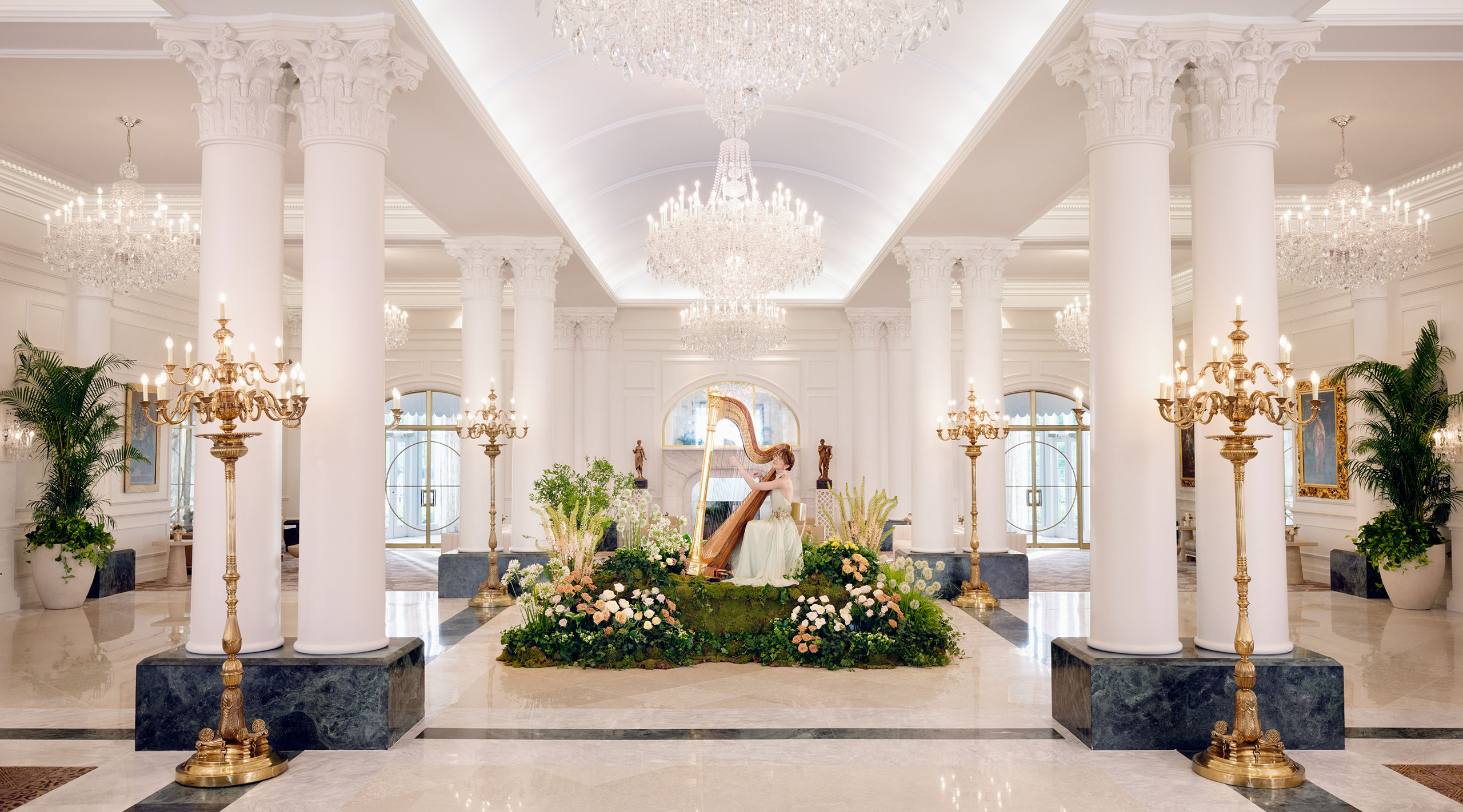 Light colors, crystal chandeliers, and large columns of the lobby of The Chateau at Nemacolin resort frame a harpist surrounded by greenery in the center of the lobby