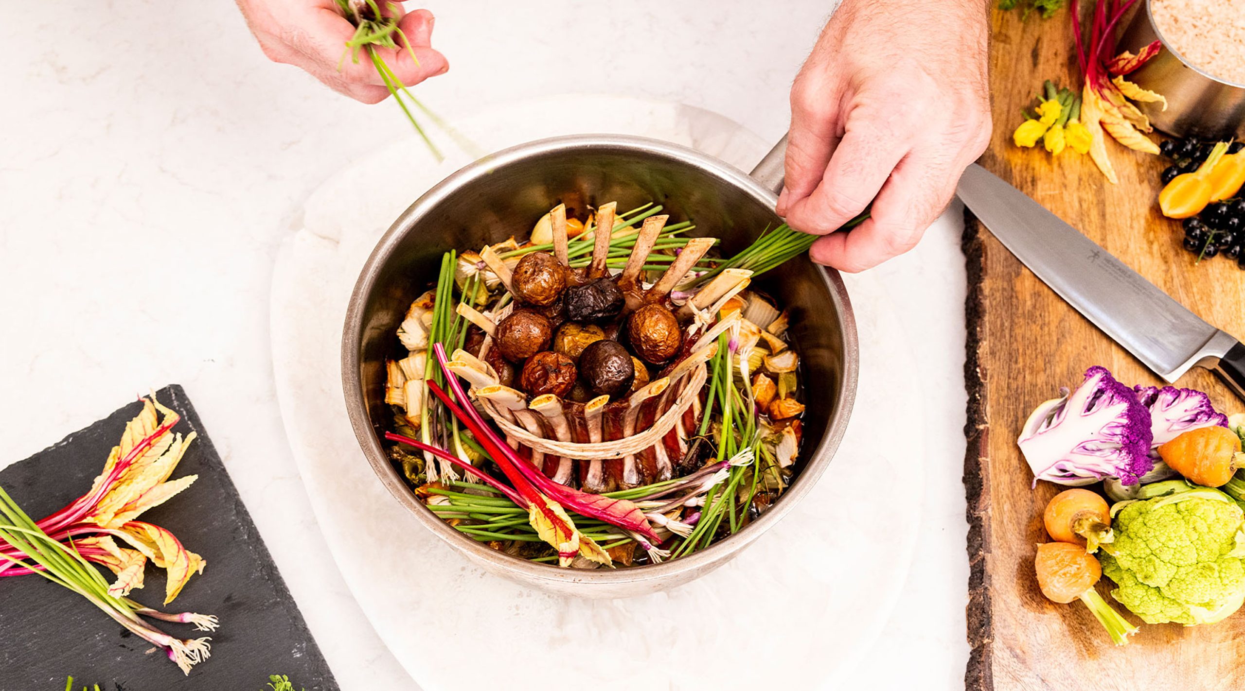 An overhead vies of a chef's hands plating a round bowl with a circular rack of meat enclosing petite potatoes and surrounded by fresh greens and vegetables