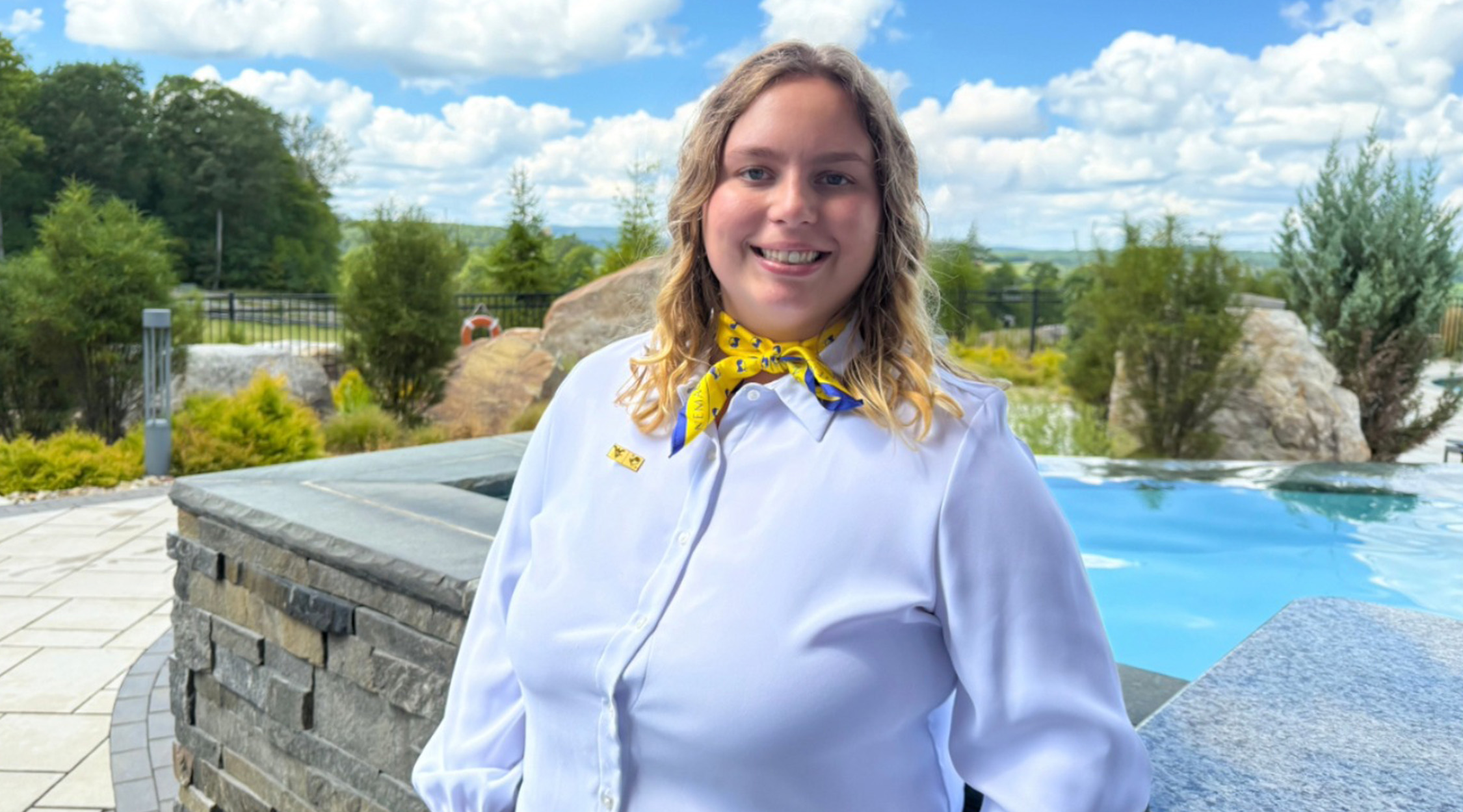A photo of Emily Eckenrode, a woman with blond hair wearing a white long-sleeved blouse and a yellow neck tie standing in front of a stone wall overlooking a pool with trees, blue sky, and clouds in the background