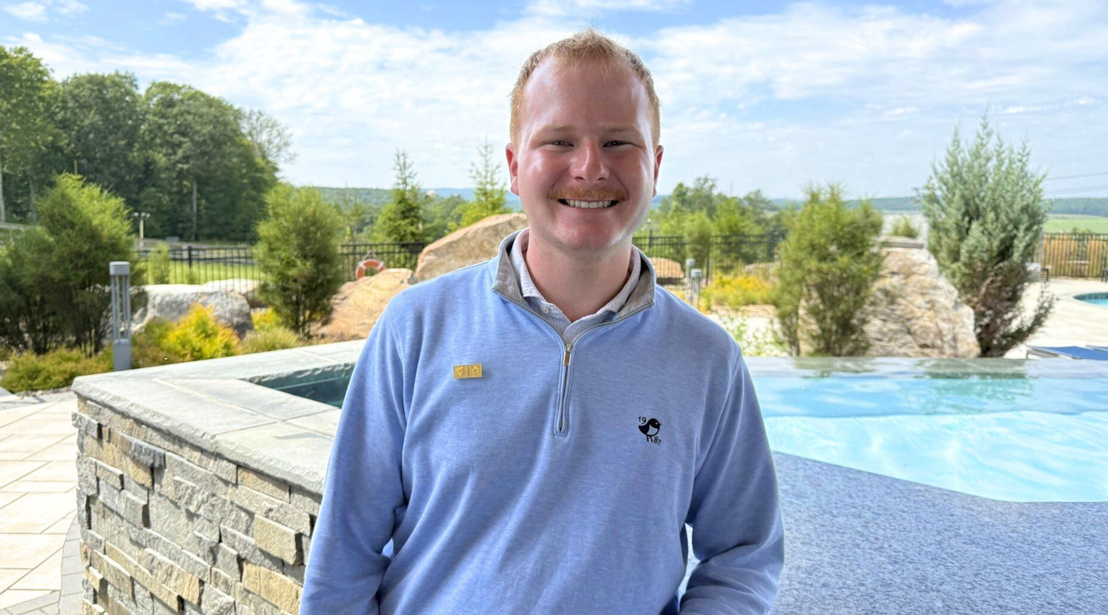 A photo of Wesley Christy, a man with short reddish-blond hair wearing a light blue long-sleeved pullover and standing in front of a stone wall overlooking a pool with trees, blue sky, and clouds in the background