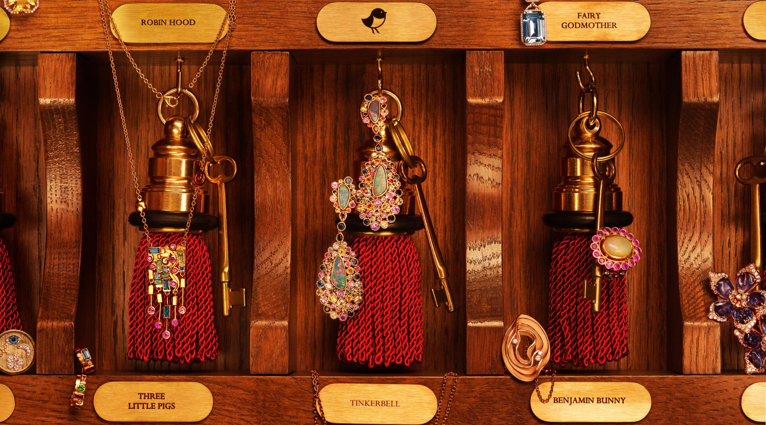 Old fashioned golden-colored skeleton keys attached to rich red tassels and adorned with multicolored jewelry pieces such as a necklace, earrings, and rings hanging in a vintage hotel keybox with visible fairytale names on gold plates above each key holder.