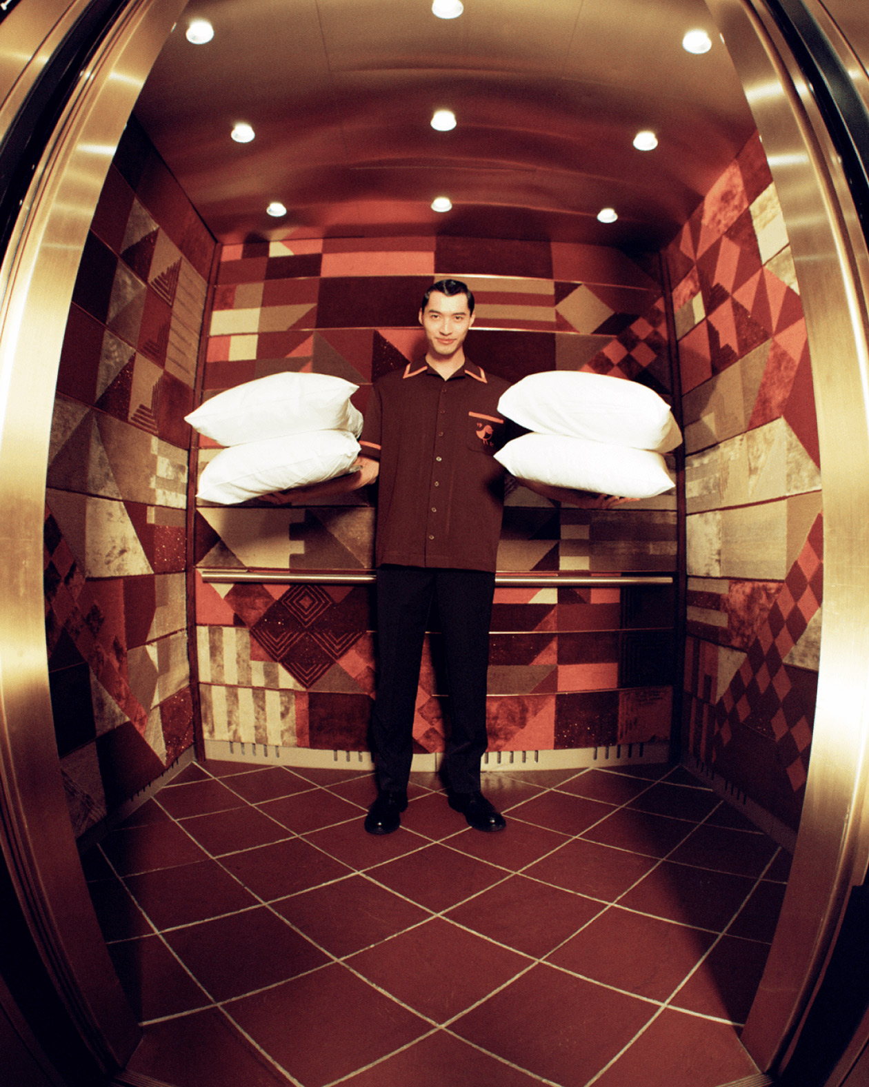 A bellhop standing in an elevator with red and patterned walls, holding a stack of white pillows. The bellhop is dressed in a dark uniform and is looking directly at the camera.