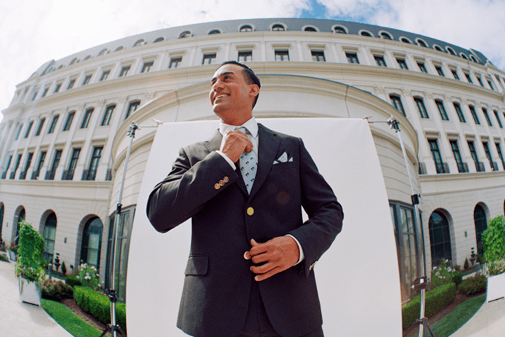 A doorman in a dark suit adjusting his tie, standing in front of a large, elegant building with a curved facade. The building has large windows and a well-maintained garden in front. The sky is clear, and the doorman is smiling confidently.